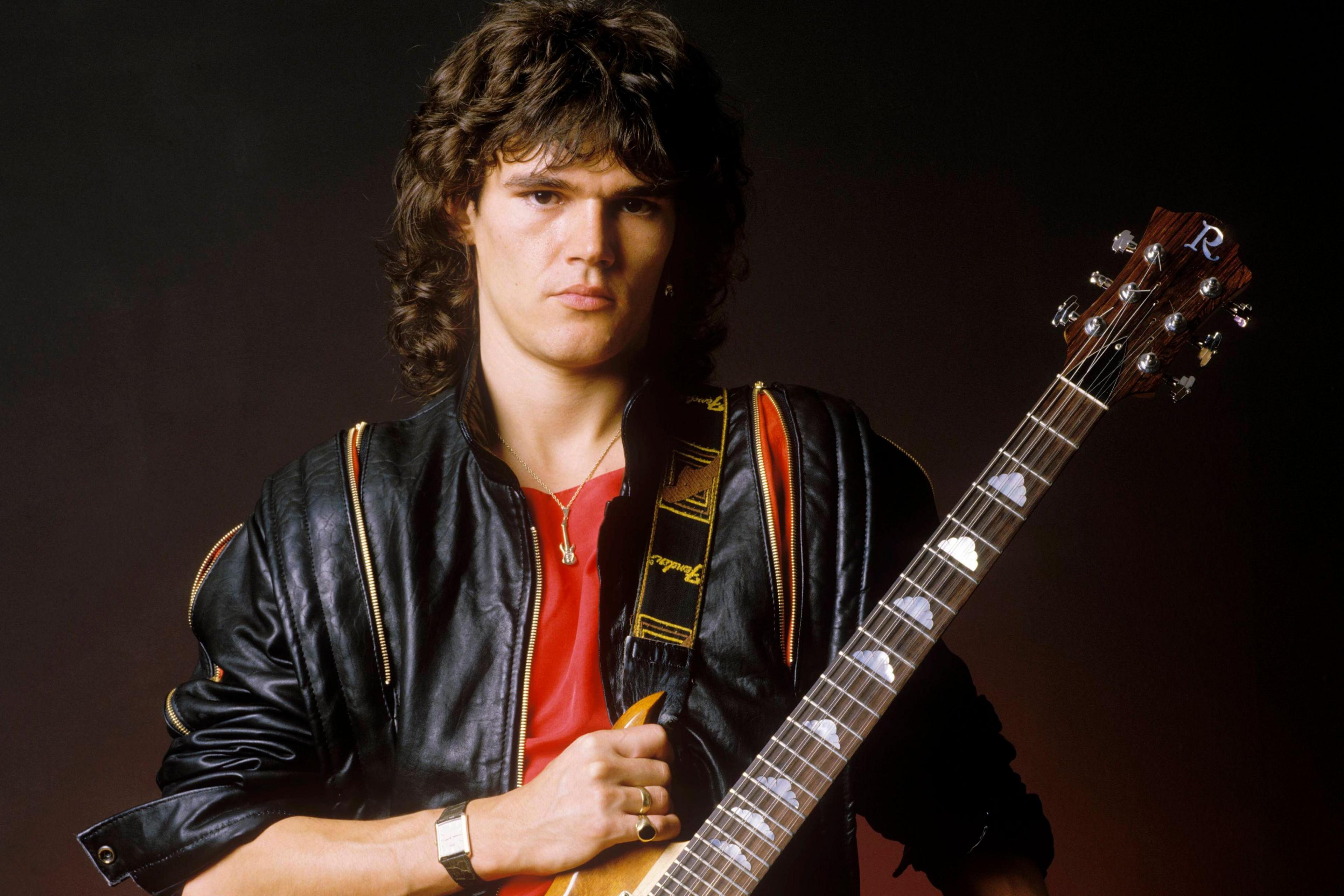 A young Robin George wears a black leather jacket and red t-shirt with a guitar slung over his left shoulder.