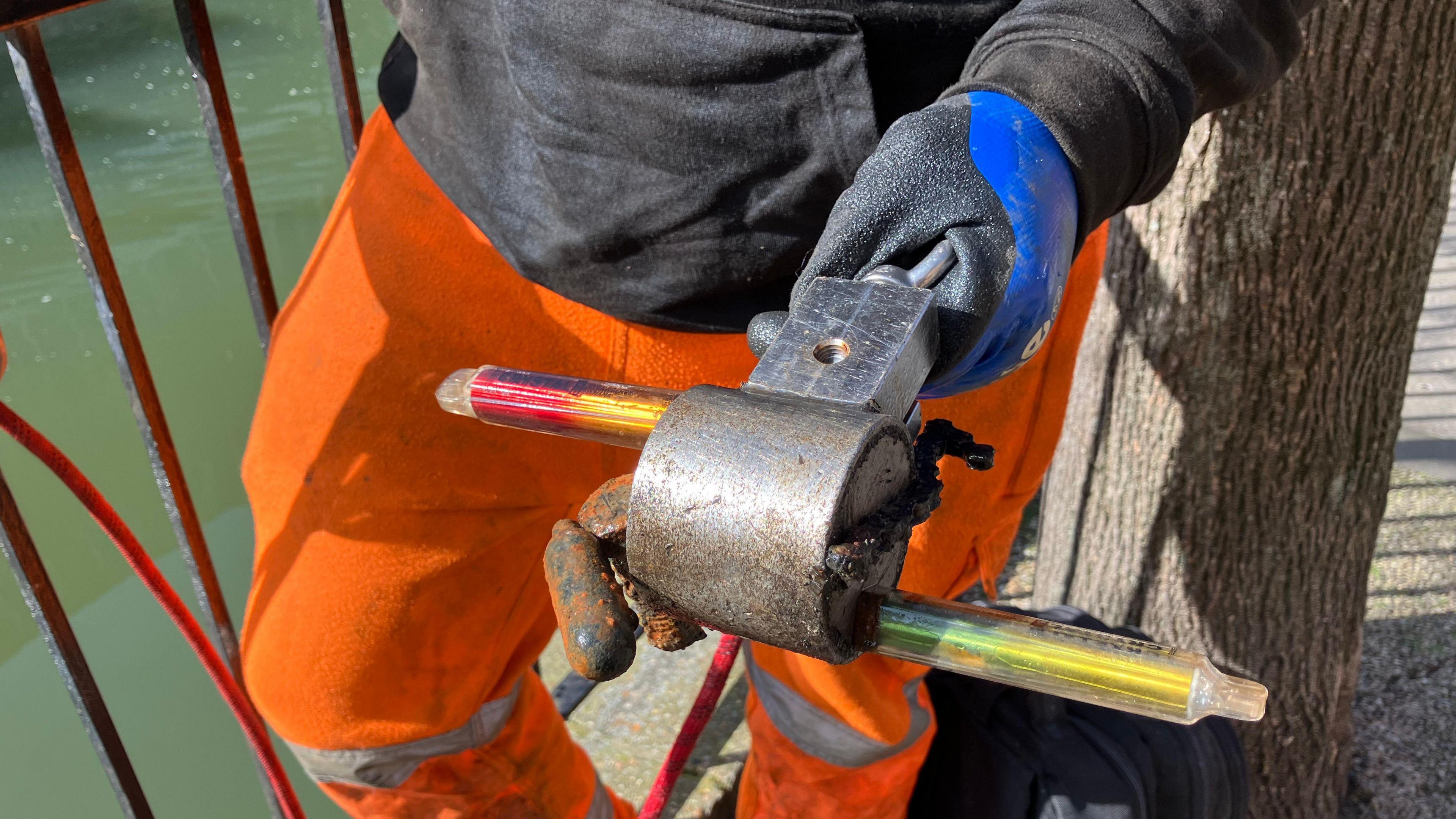 A metal object with two disposable vapes stuck to it, in the background is a gloved man with bright orange trousers.