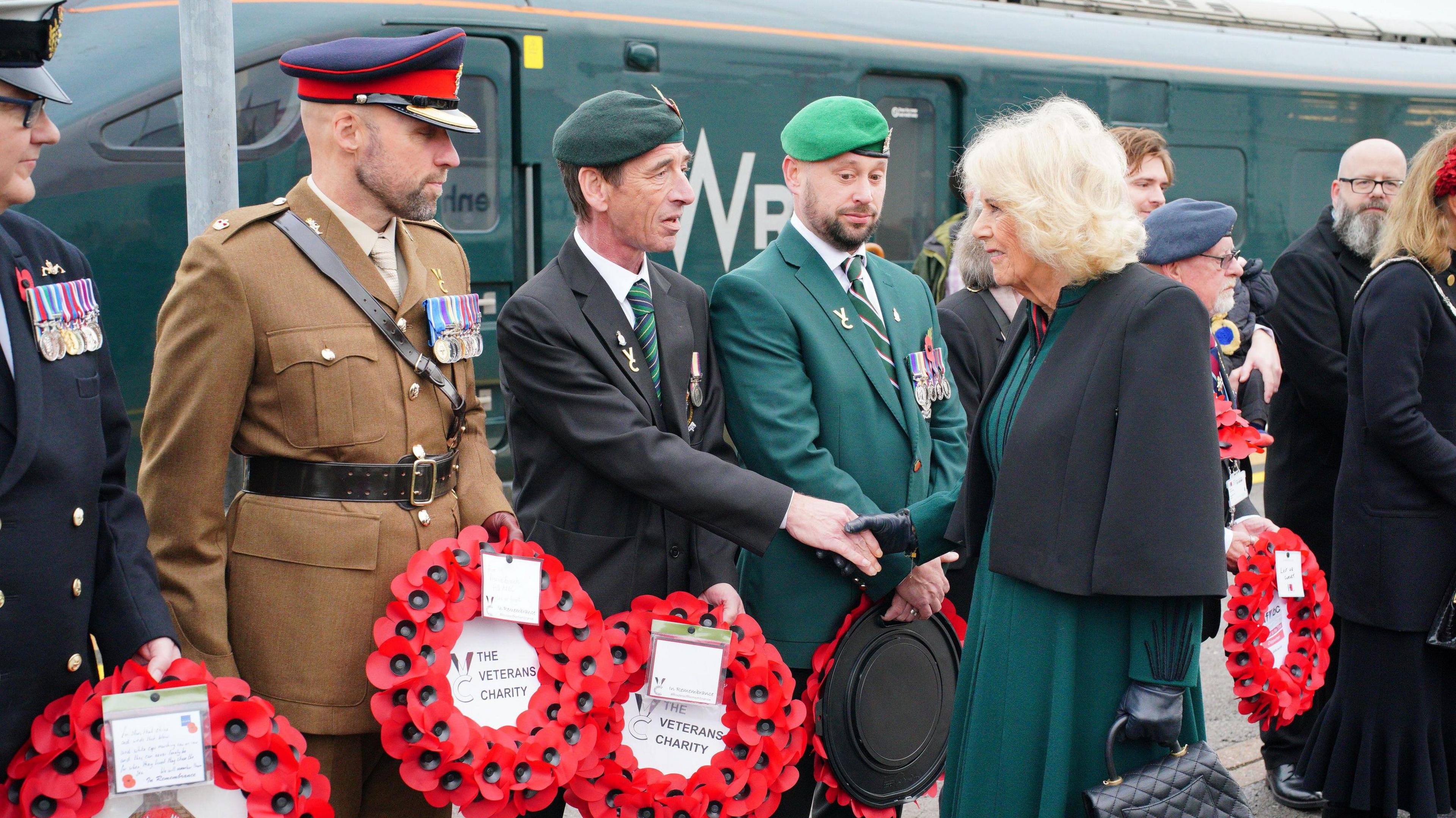 The Queen shakes hand with a smartly dressed veteran, who stands in a line of other veterans holding wreaths and many in uniform. All are in front of a dark green GWR train.