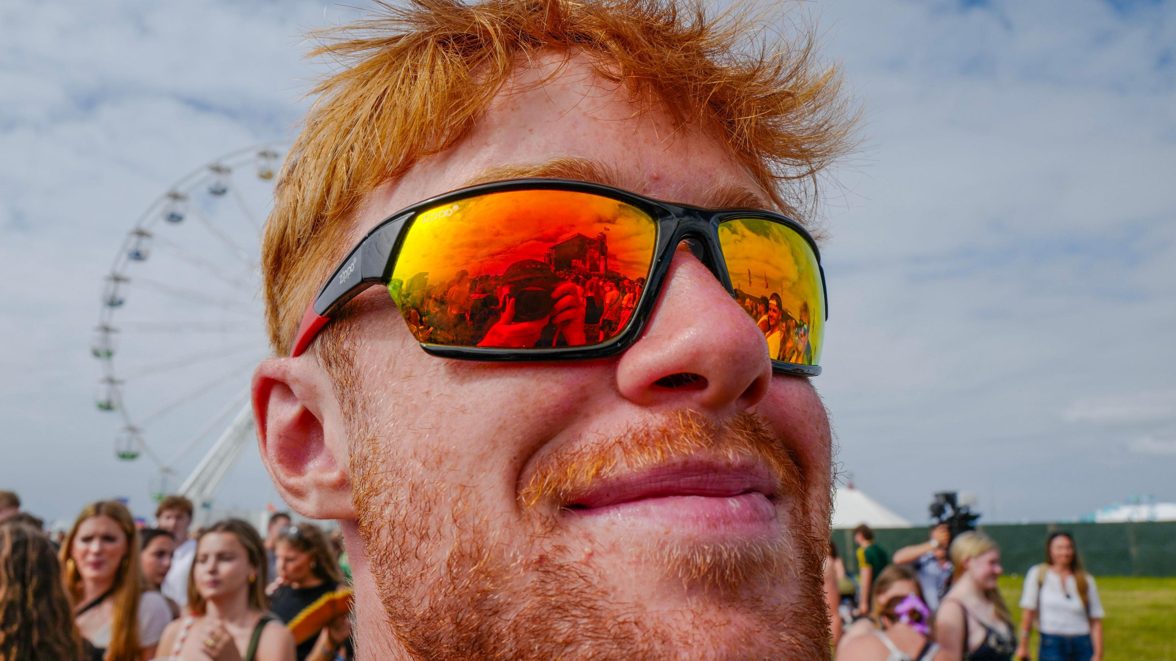 A man with bright ginger hair and stubble is seen wearing reflective orange sunglasses showing a crowd in front of him. There is a ferris wheel behind him.