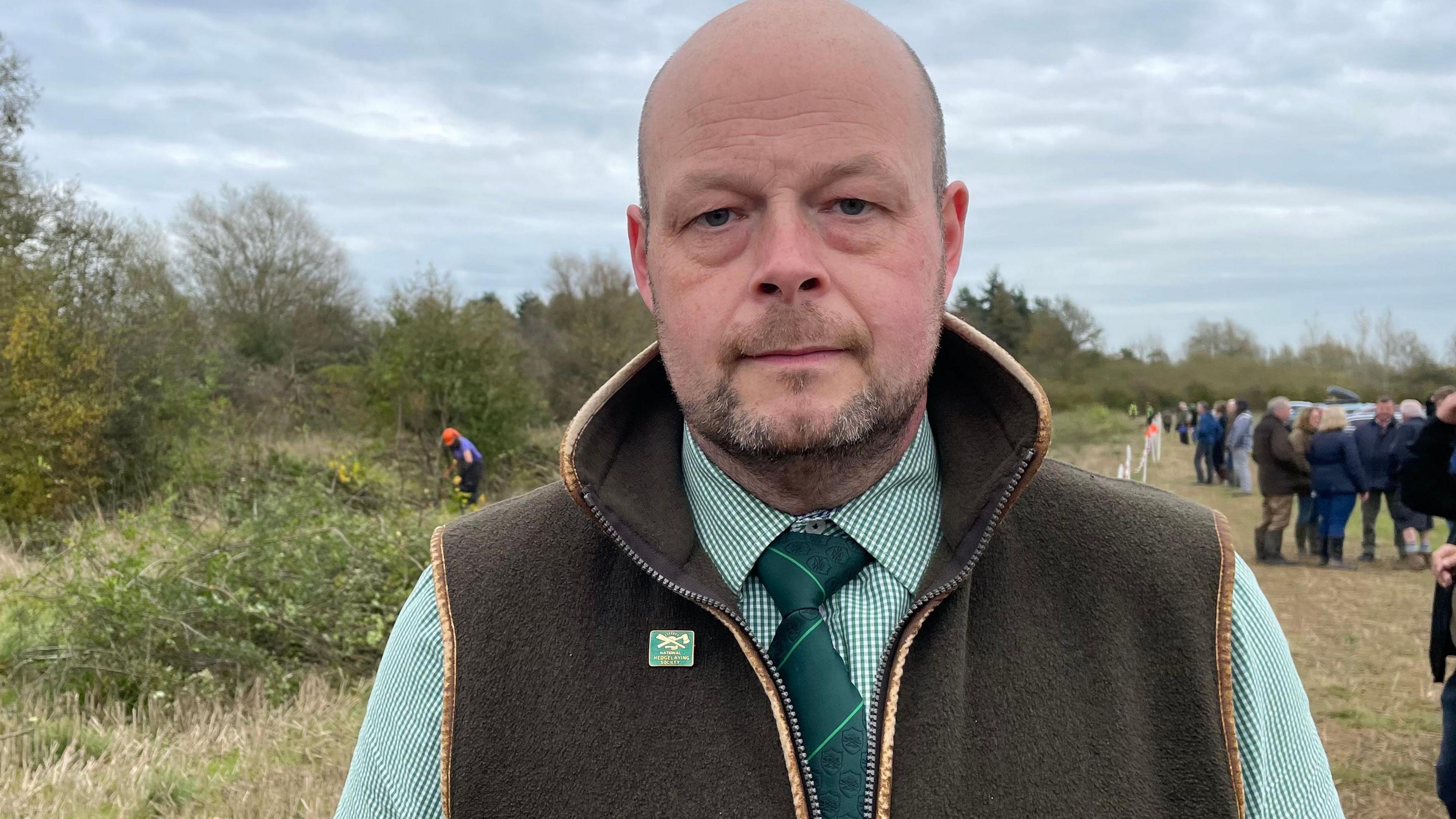 A man with a balding head, wearing a green check shirt and tie, with a gillet on top. He stands in front of hedgelayers and spectators at the competition.