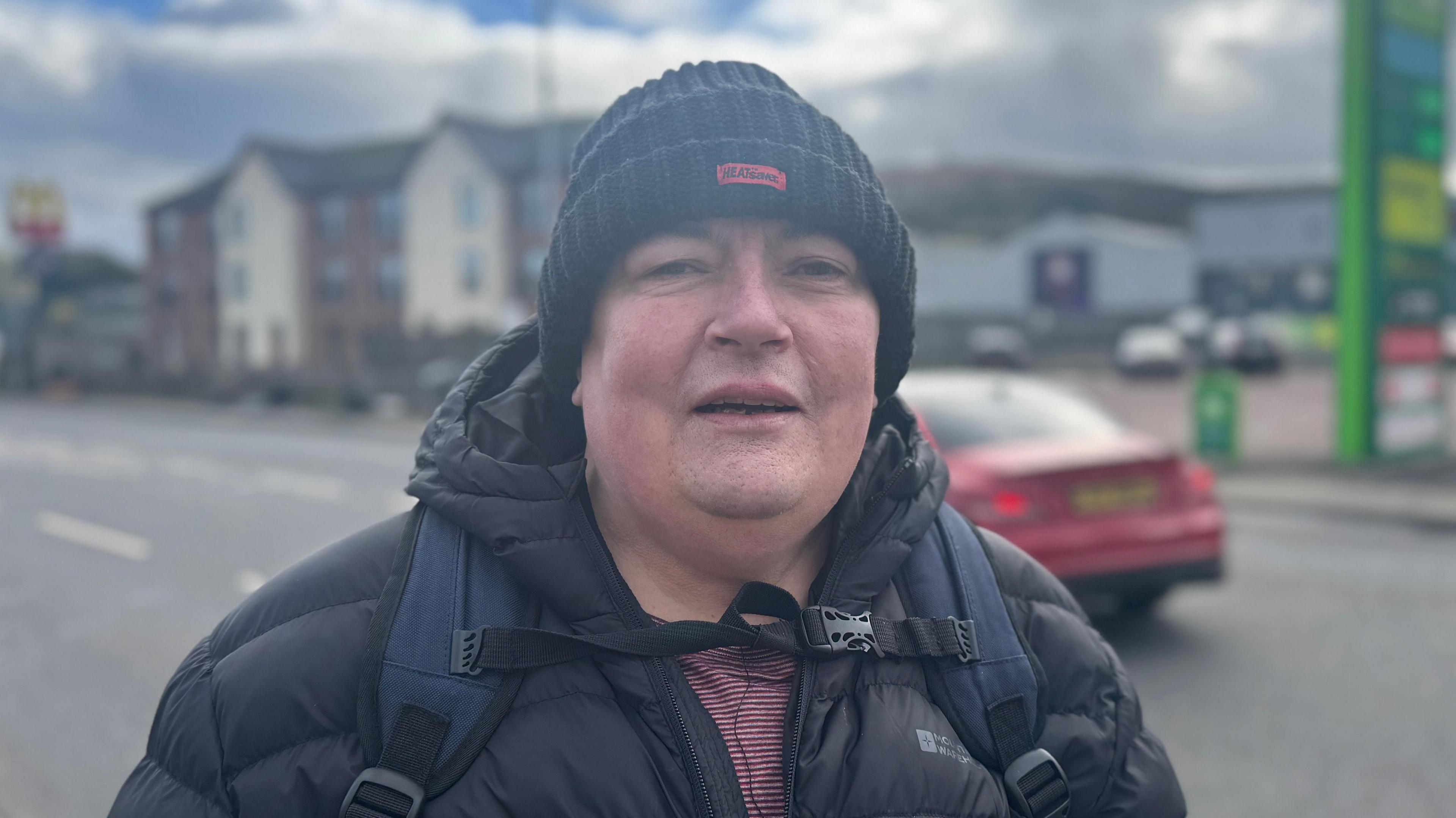 Gerald McGarva. A man stands with his mouth open while looking at the camera. He is wearing a ribbed, black beanie, a black puffer raincoat and a navy backpack. He is standing in front of a road as a red car drives by in the background.