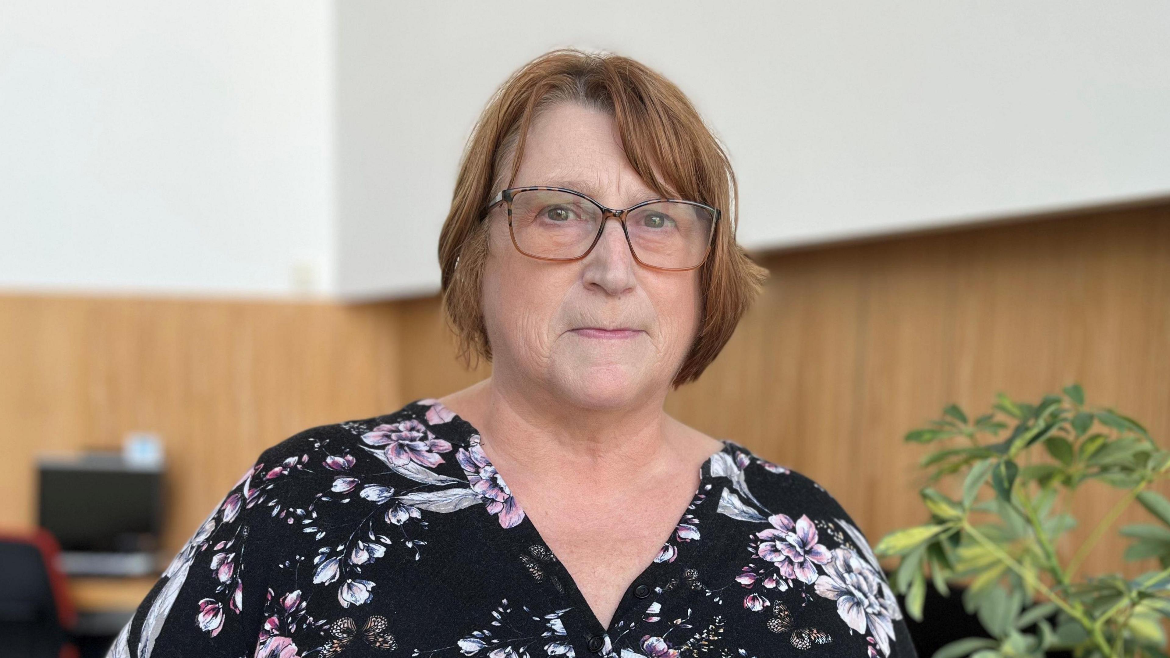 anne-marie is inside looking directly at the camera. She wears glasses and has brown hair. She is wearing a flowery dress