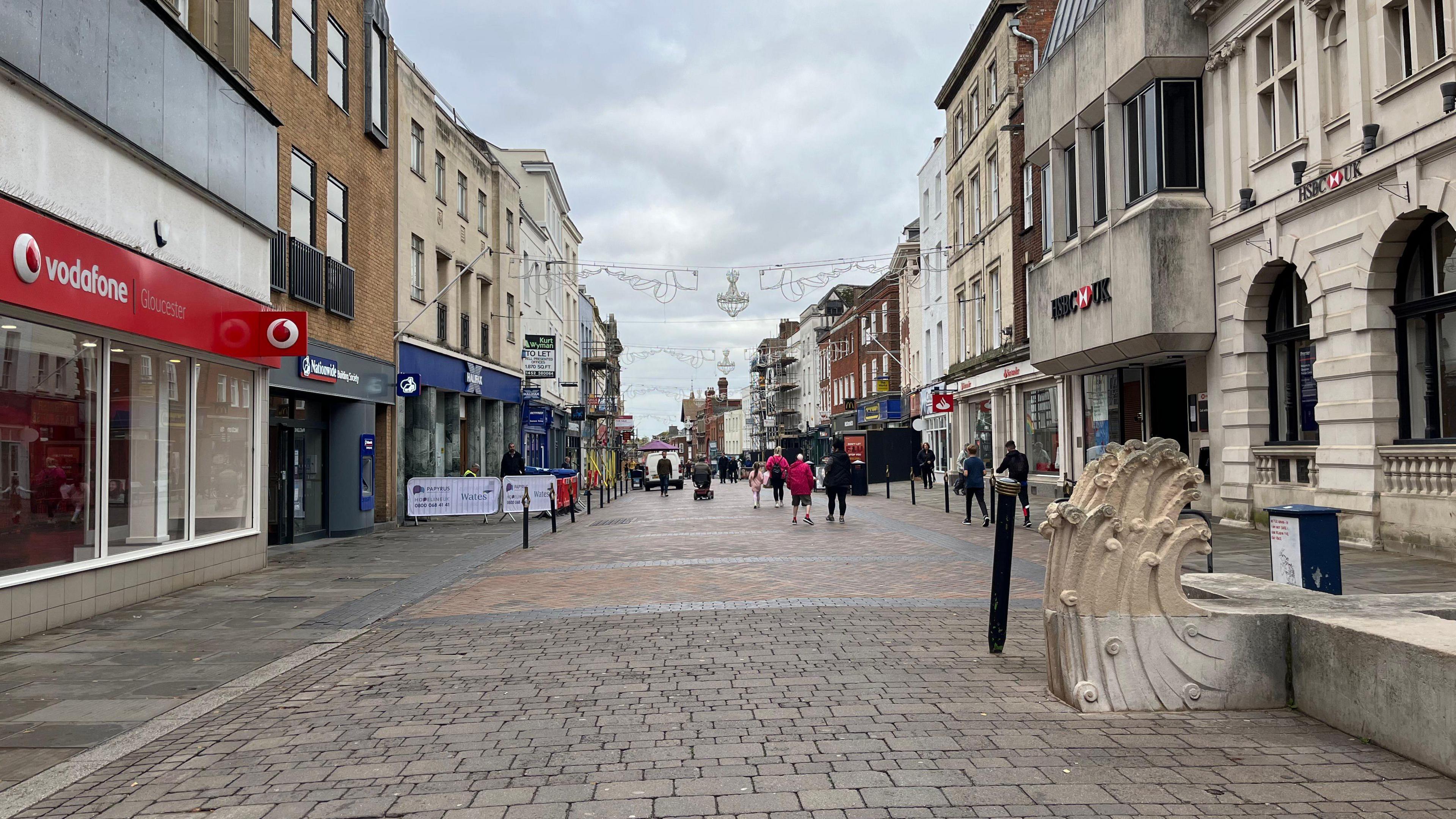 A photo showing Westgate Street in Gloucester. The street is cobbled with shop fronts on either side and shopping shown walking along