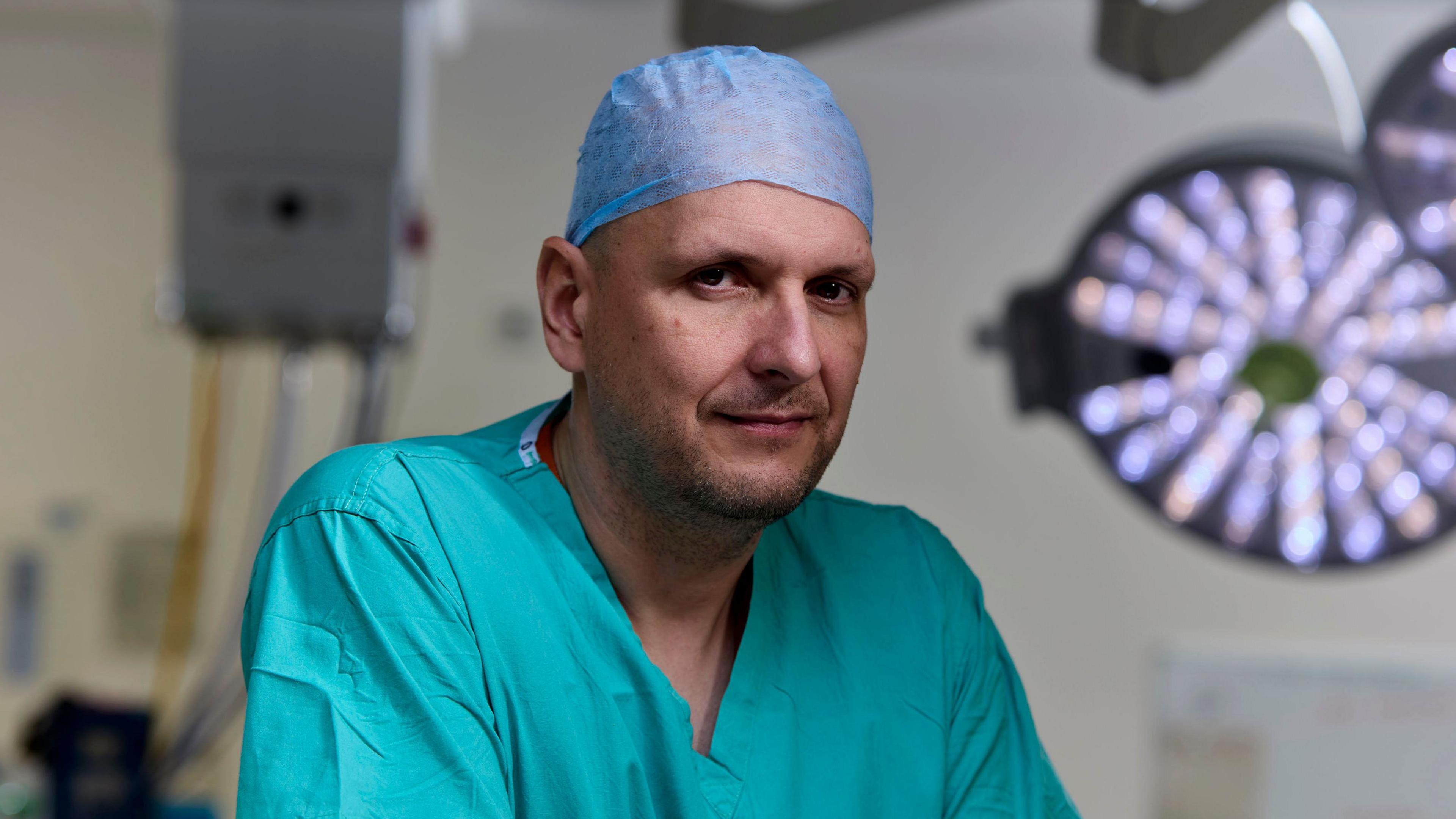 Consultant neurosurgeon Drahoslav Sokol leans on a surface in the operating theatre, wearing turquoise blue scrums and a pale blue theatre cap, the circular bright light behind him and monitors in the background