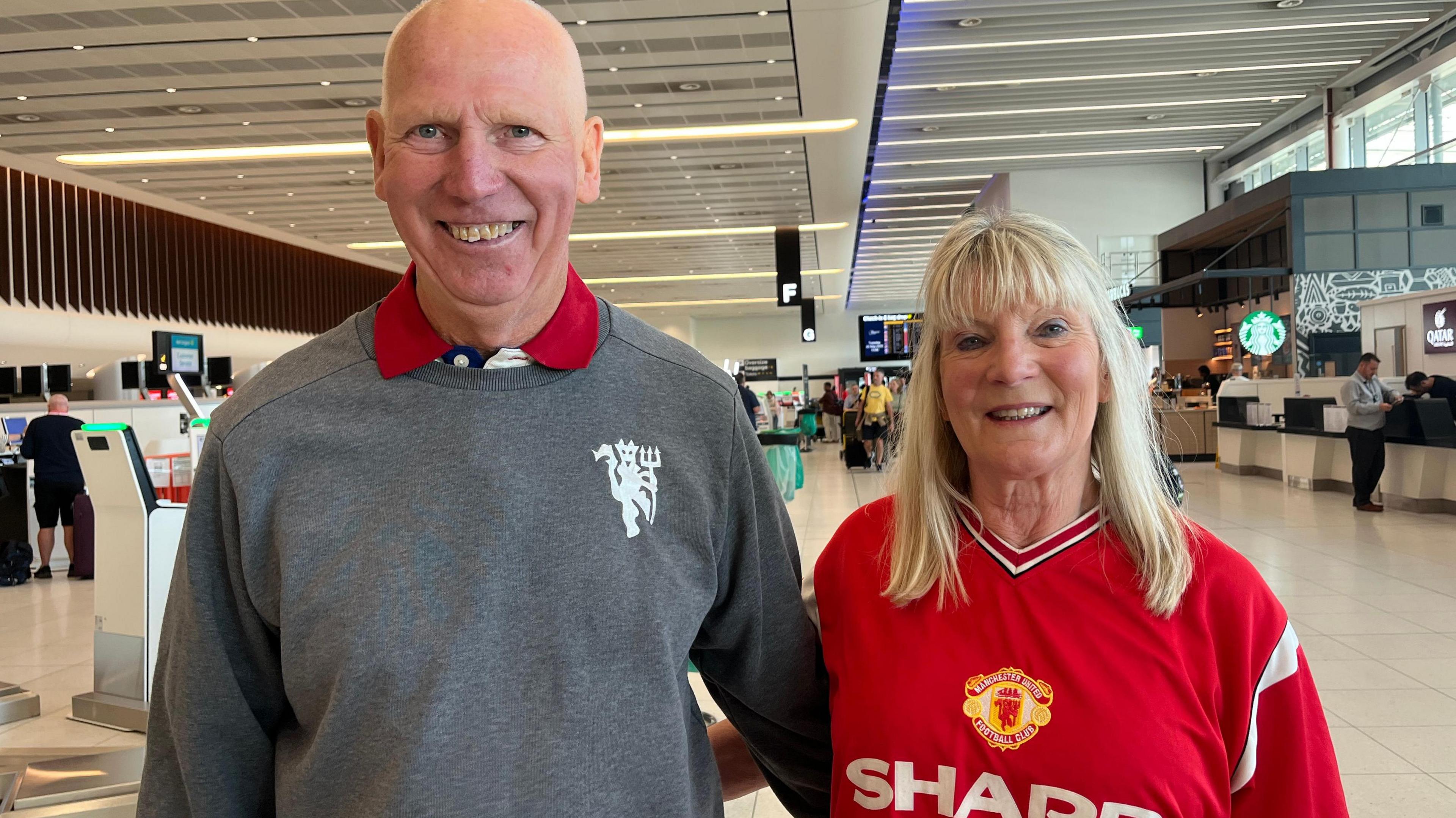 Jim is bald and is wearing a grey crew neck sweater with a white Manchester United crest while his wife has blonde hair and is wearing a replica Manchester United shirt.