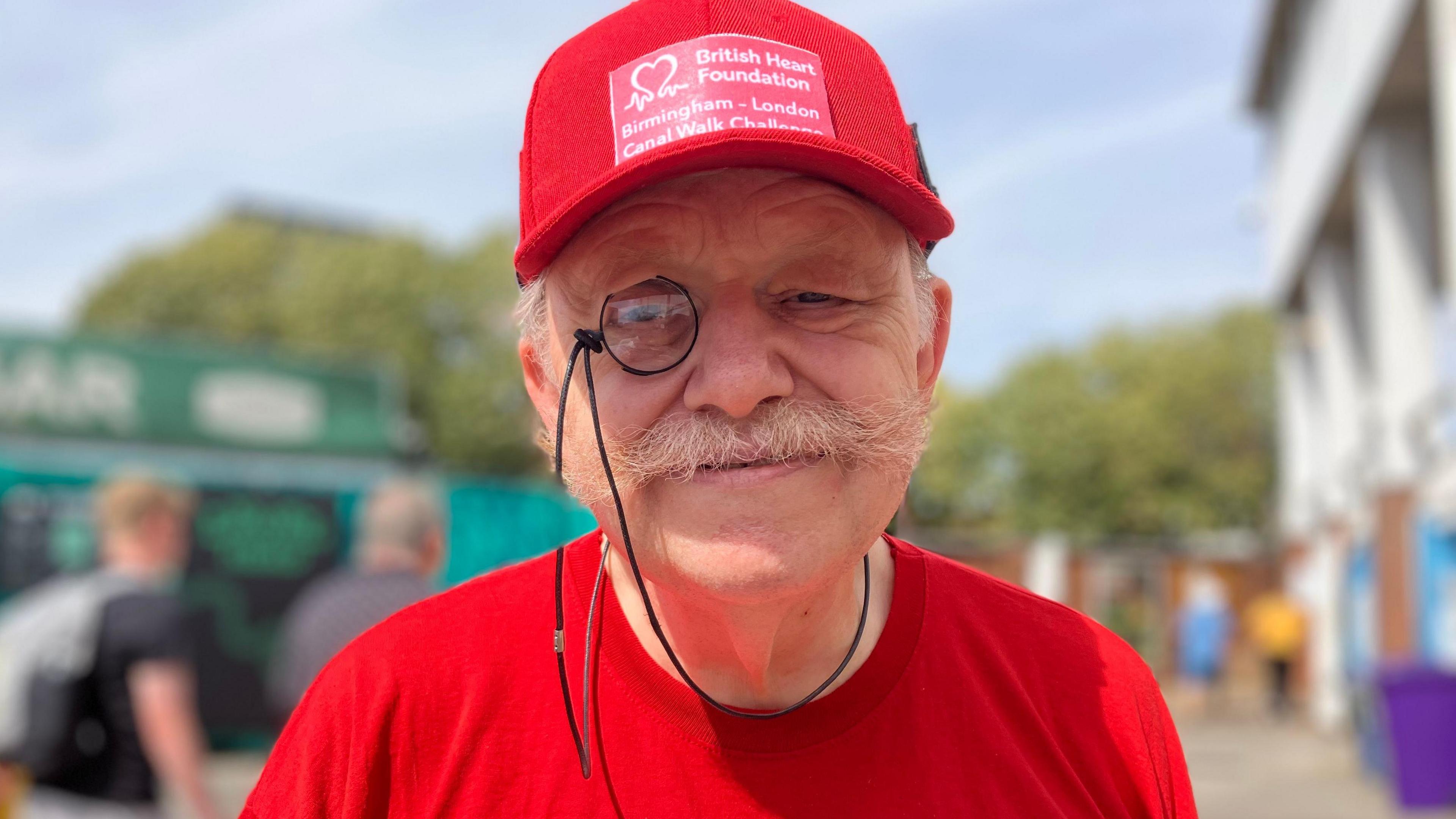 David Tomlinson smiles and looks into the camera. He is wearing a red cap and a red t-shirt. He has a monocle and a grey moustache. In the background is one of the stands at Trent Bridge Cricket Ground and some other spectators