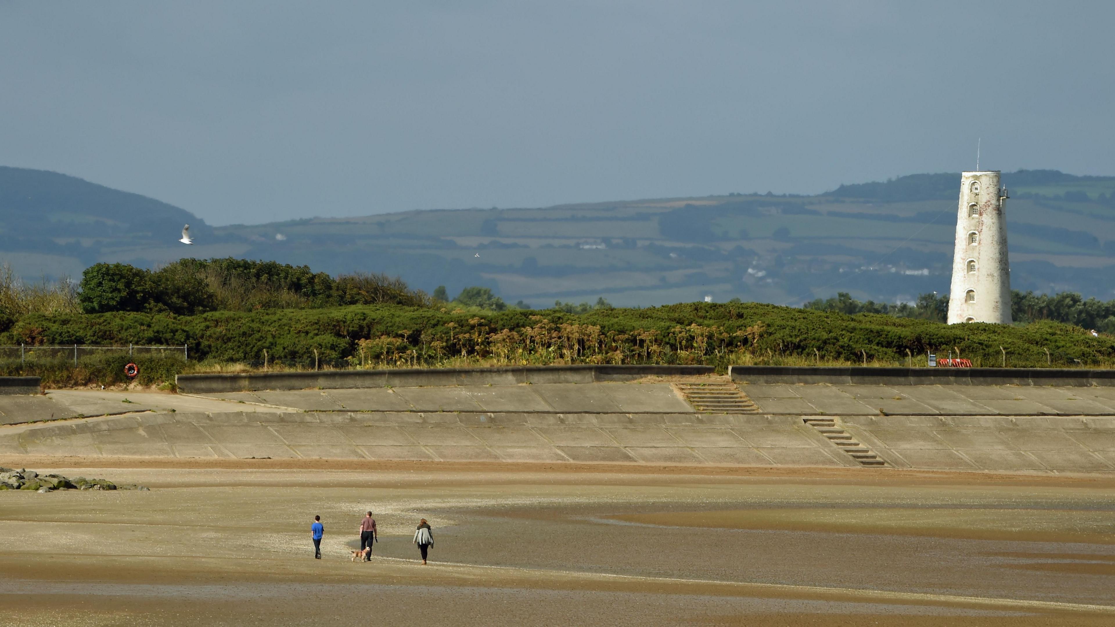 Stock image of three people walking on the beach with dogs. Leasowe Lighthouse, which is a white building, can be seen perched on higher ground on the right with hills in the background.