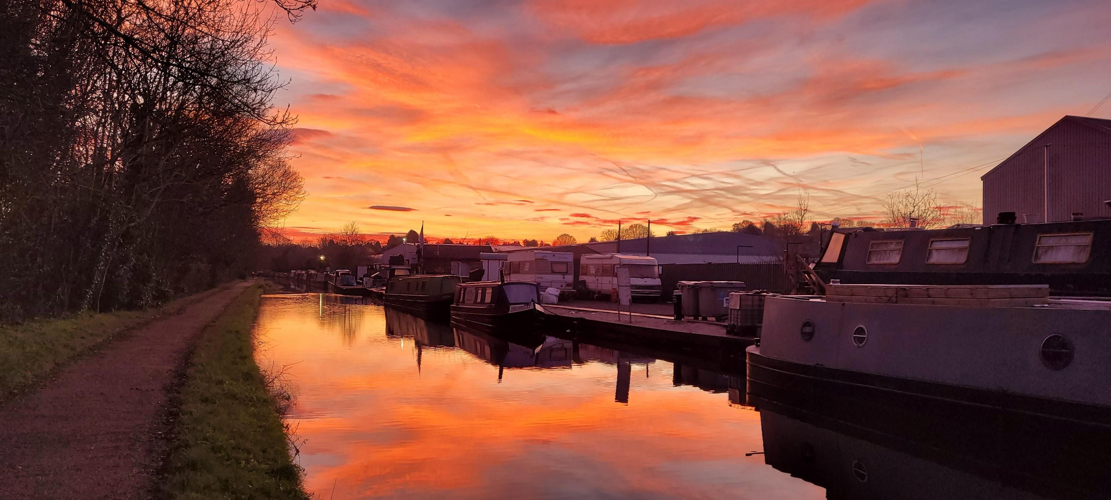 Various shades of orange dominate an otherwise pale-blue sky over a canal. Three moored narrowboats line up along one side, and behind them two motor homes stand in a fenced area with industrial buildings behind. 