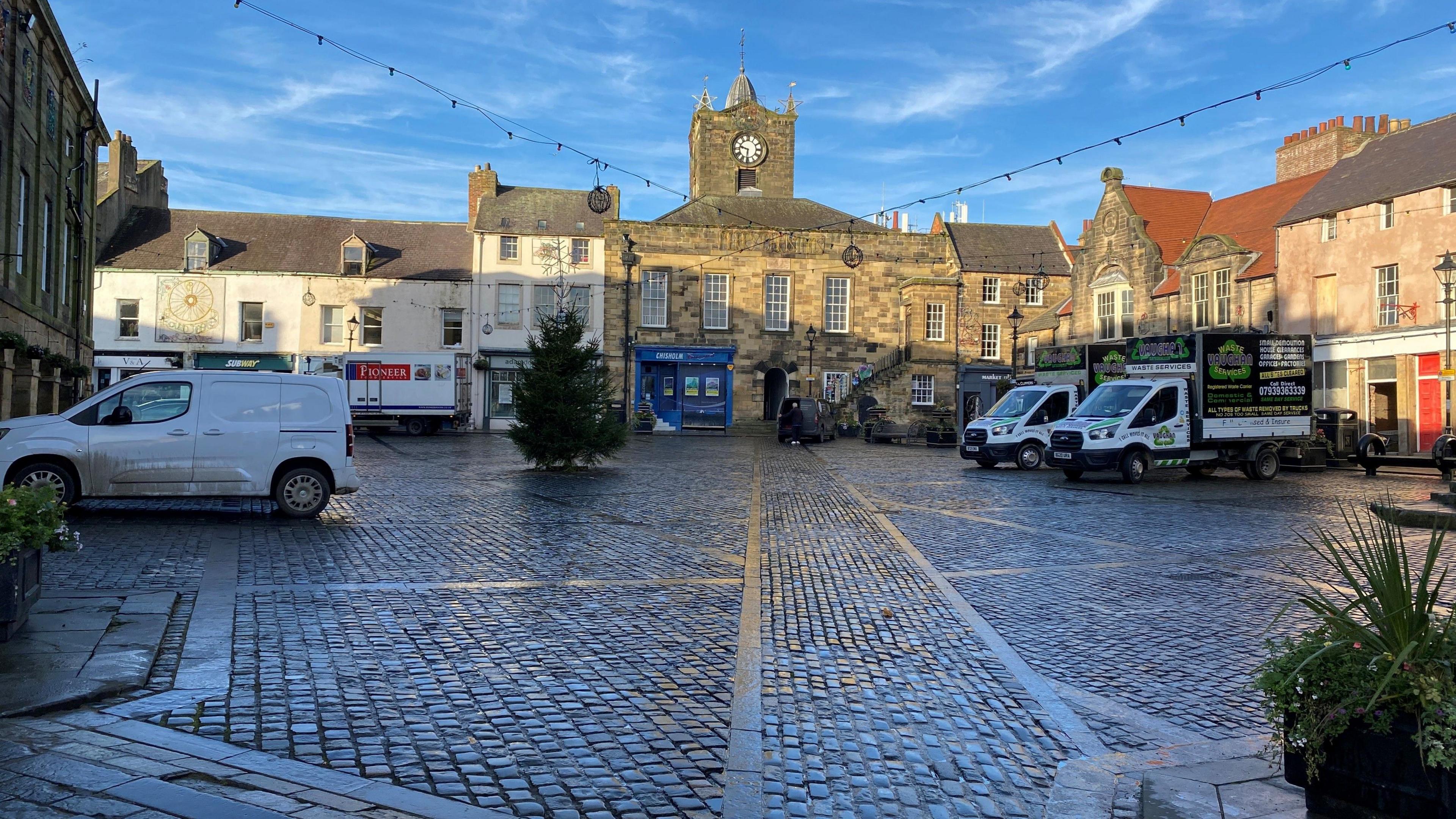 A small Christmas tree in the centre of Alnwick's market place with a number of vehicles parked around it. 