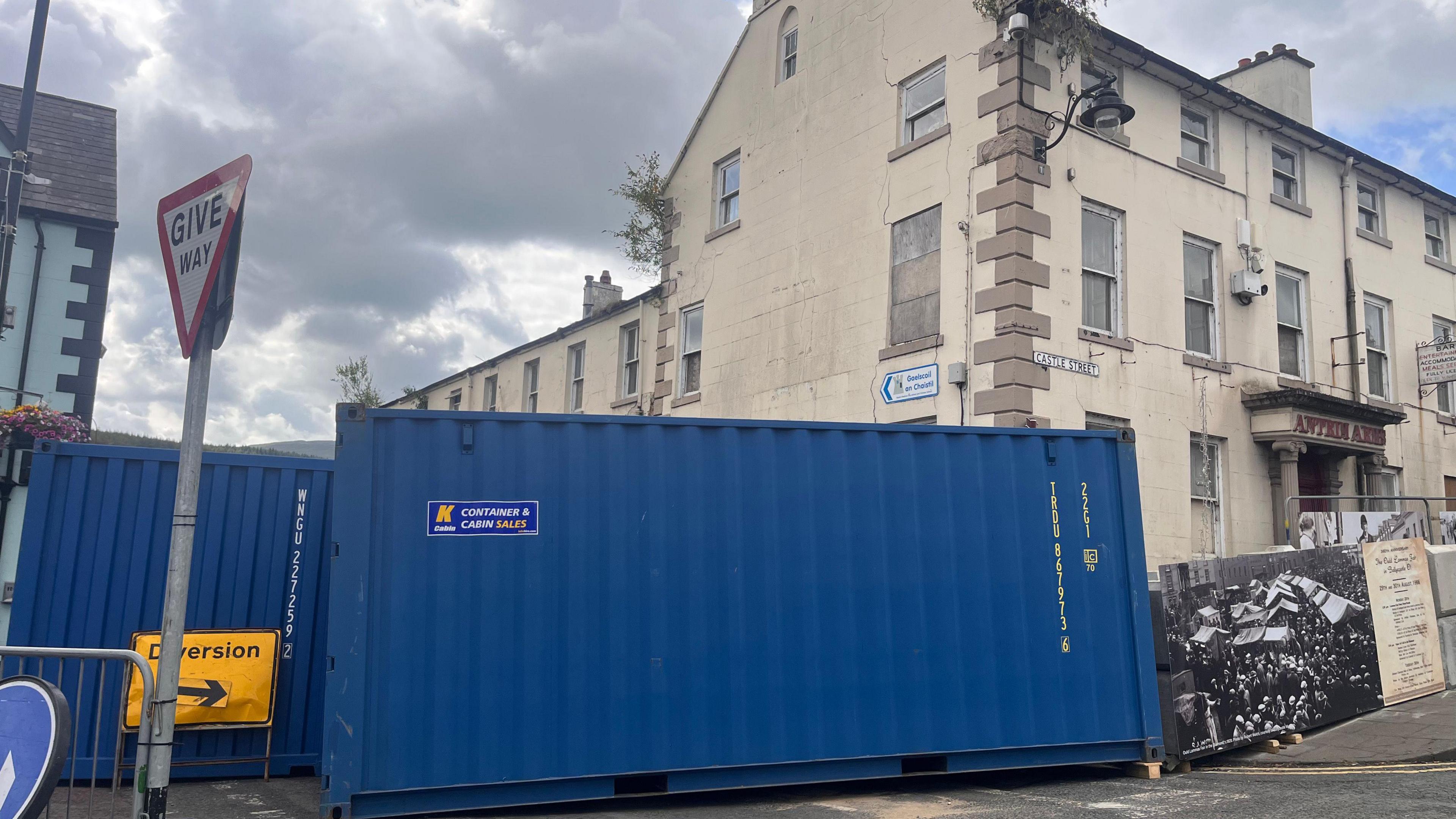Blue shipping containers outside a derelict hotel. The sky is cloudy. A yellow sign reads 'diversion'.