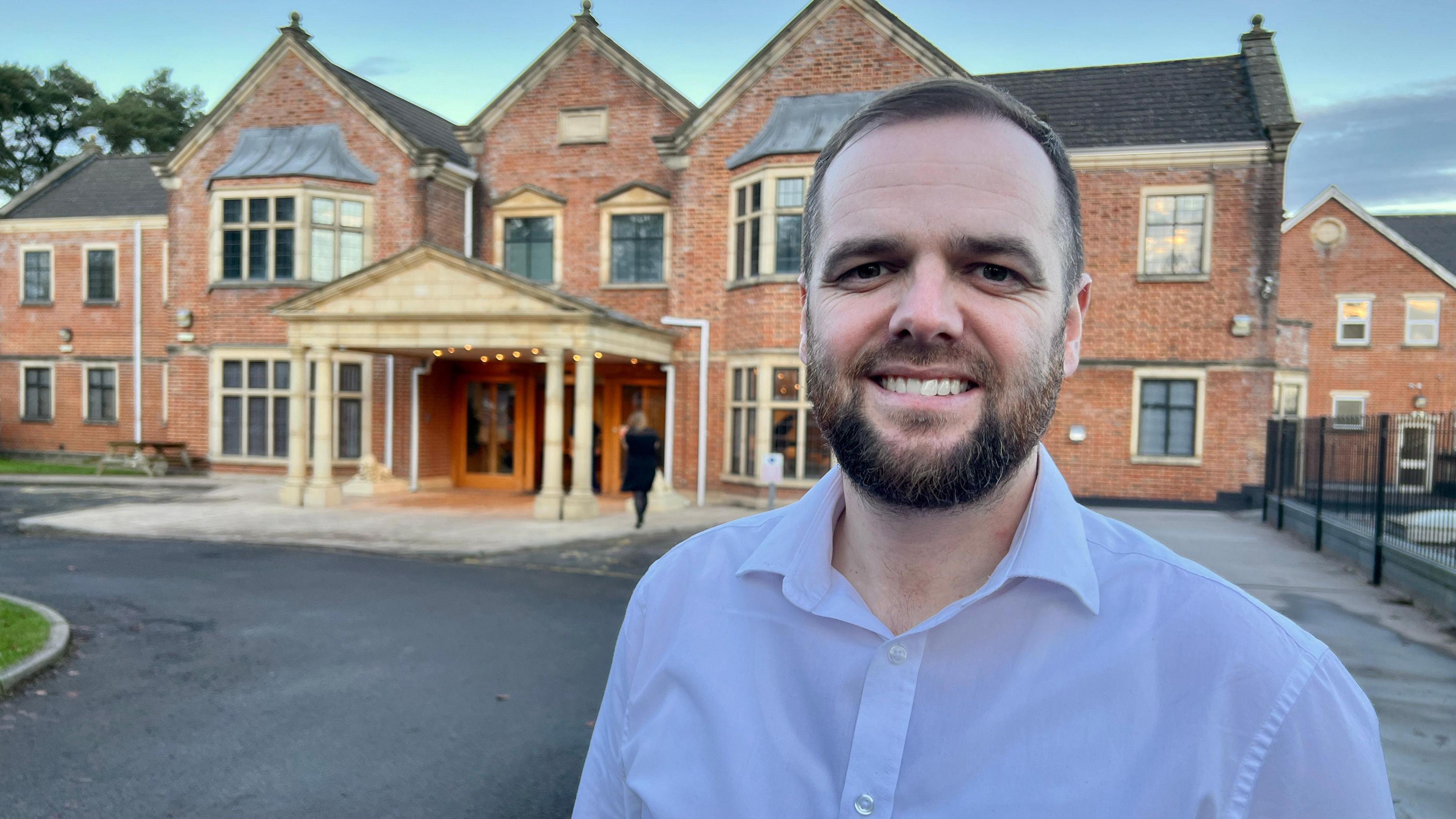 A man with a beard wearing a white shirt smiling at the camera. Behind him is a hall-type building