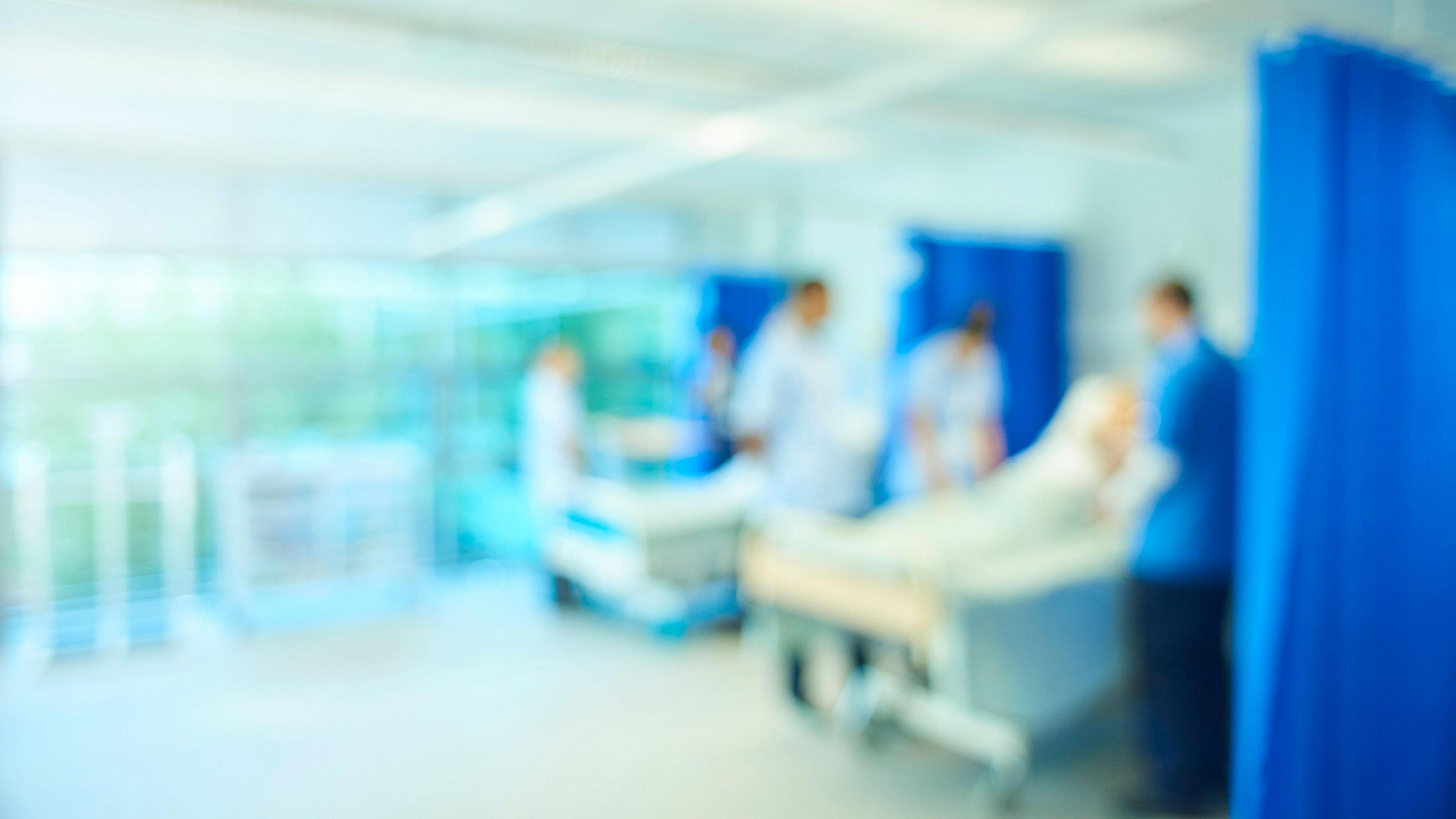 A blurred image of what appears to be a hospital ward with medical staff in blue uniforms moving between beds. The image is intentionally out of focus, giving a sense of constant movement and activity.