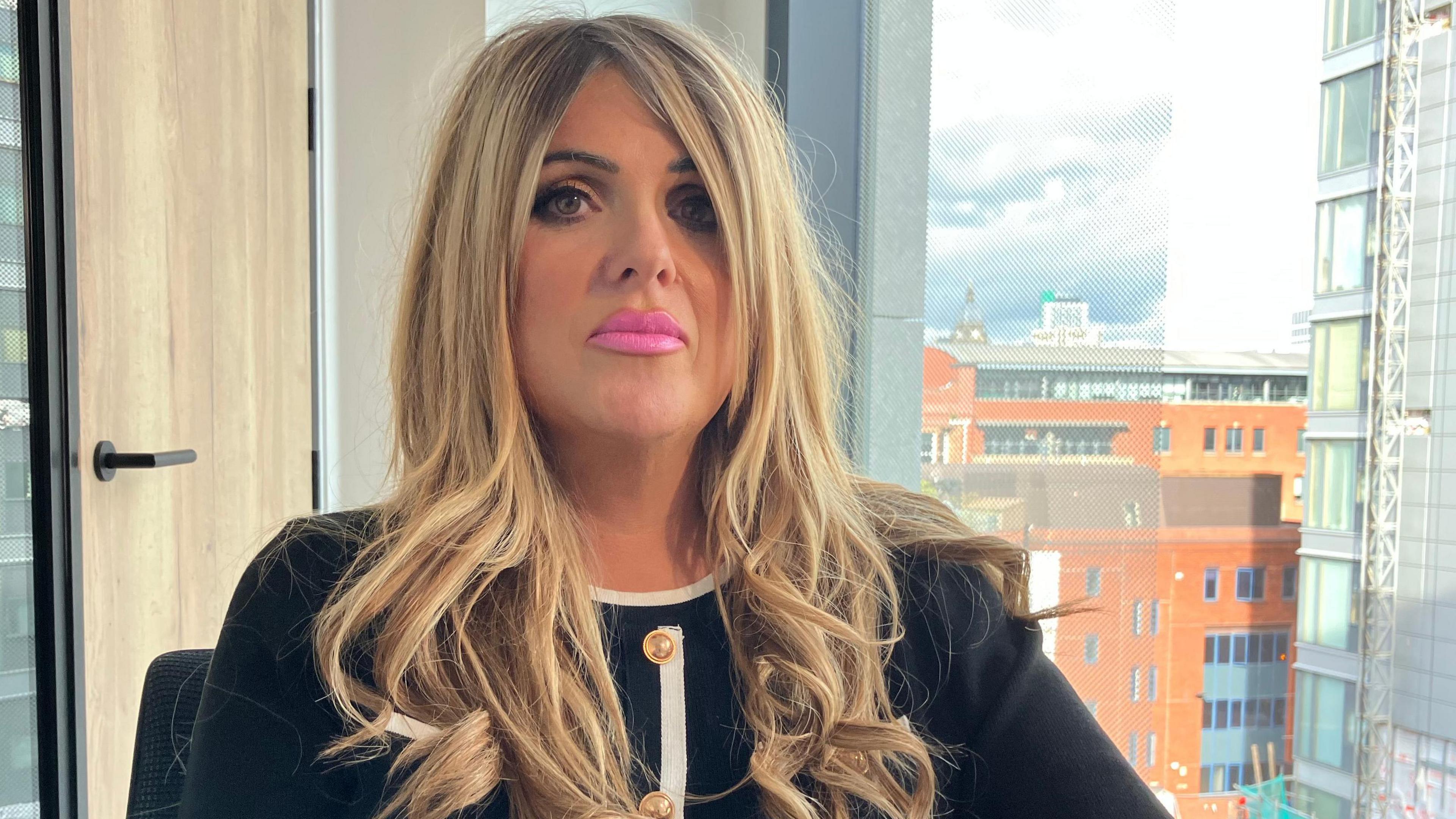 A woman with blonde-brown hair. The ends are curled. She wears a white long-sleeved shirt or blazer, with white hems. She has pink lipstick on and looks at the lens with a neutral expression.