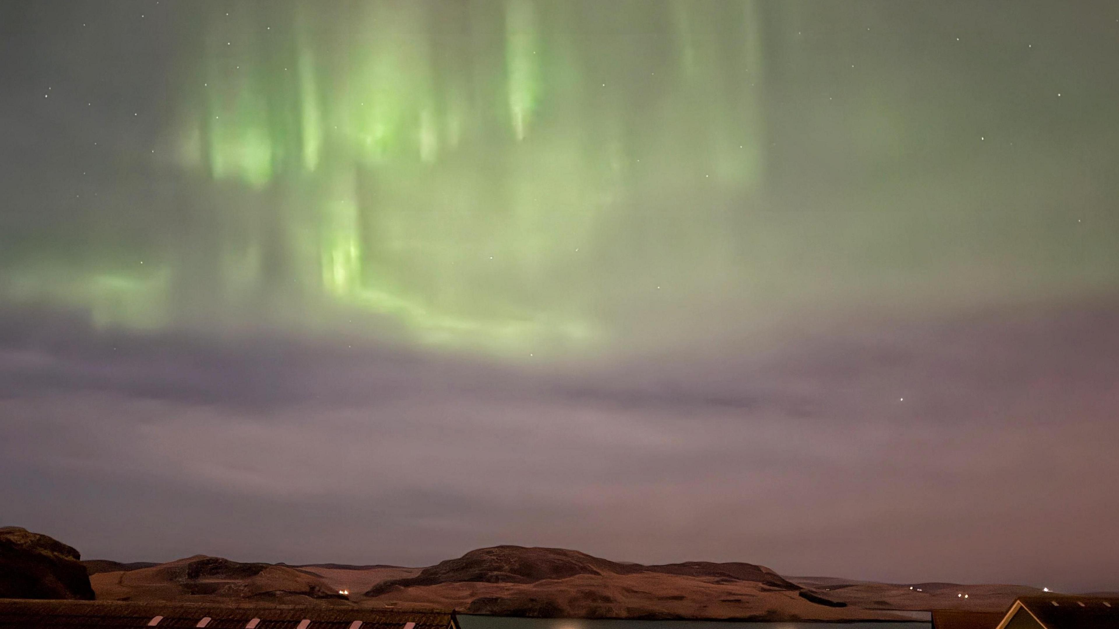 Bright green aurora in a purple night sky above houses and hills