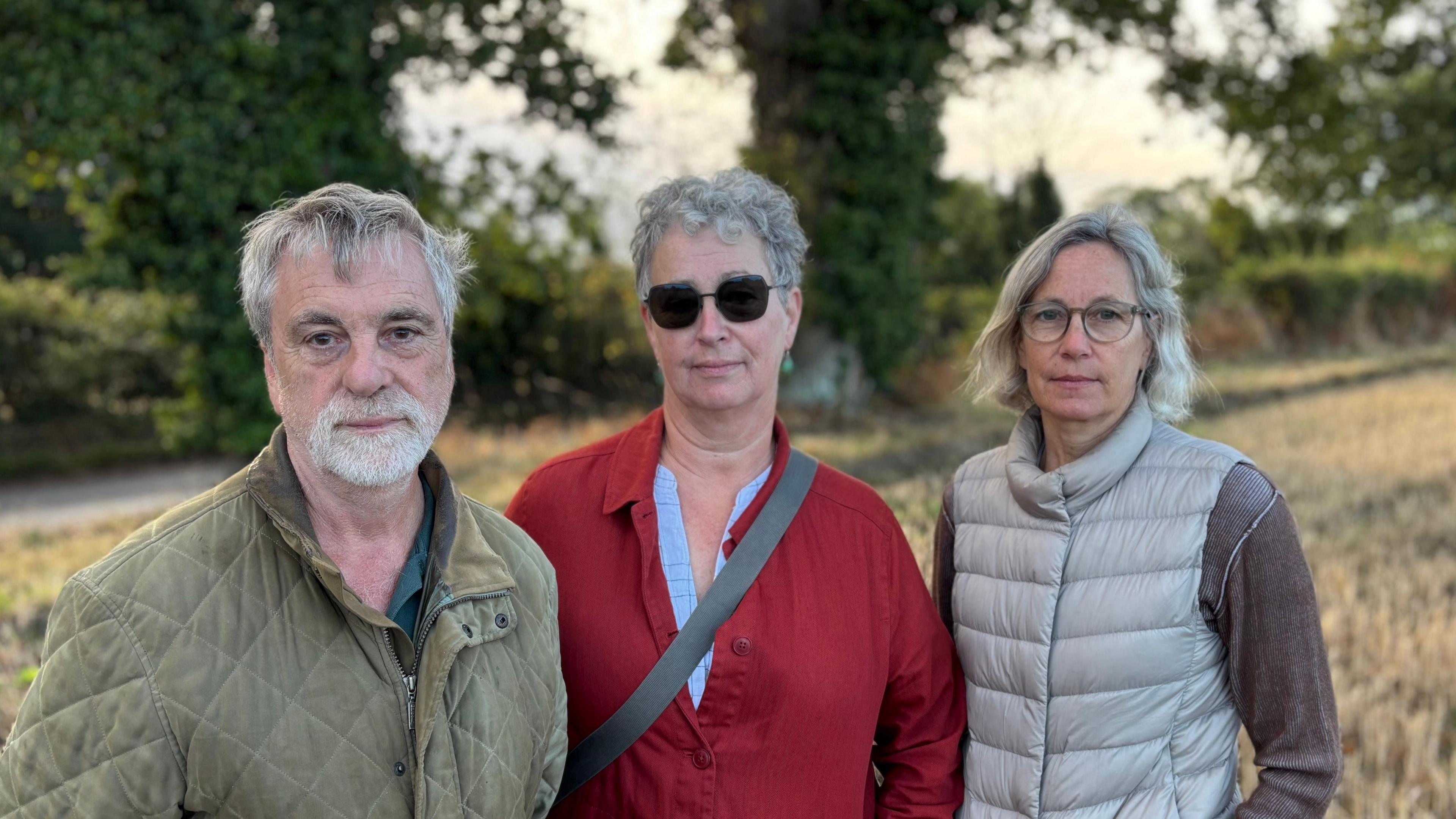 A man and two women standing next two each other in a field and in front of some trees. The man is wearing a green jacket, the middle woman is wearing a red jacket with dark glasses, and the second woman, pictured on the right, is wearing a grey Gillet and glasses. They are all looking into the camera.