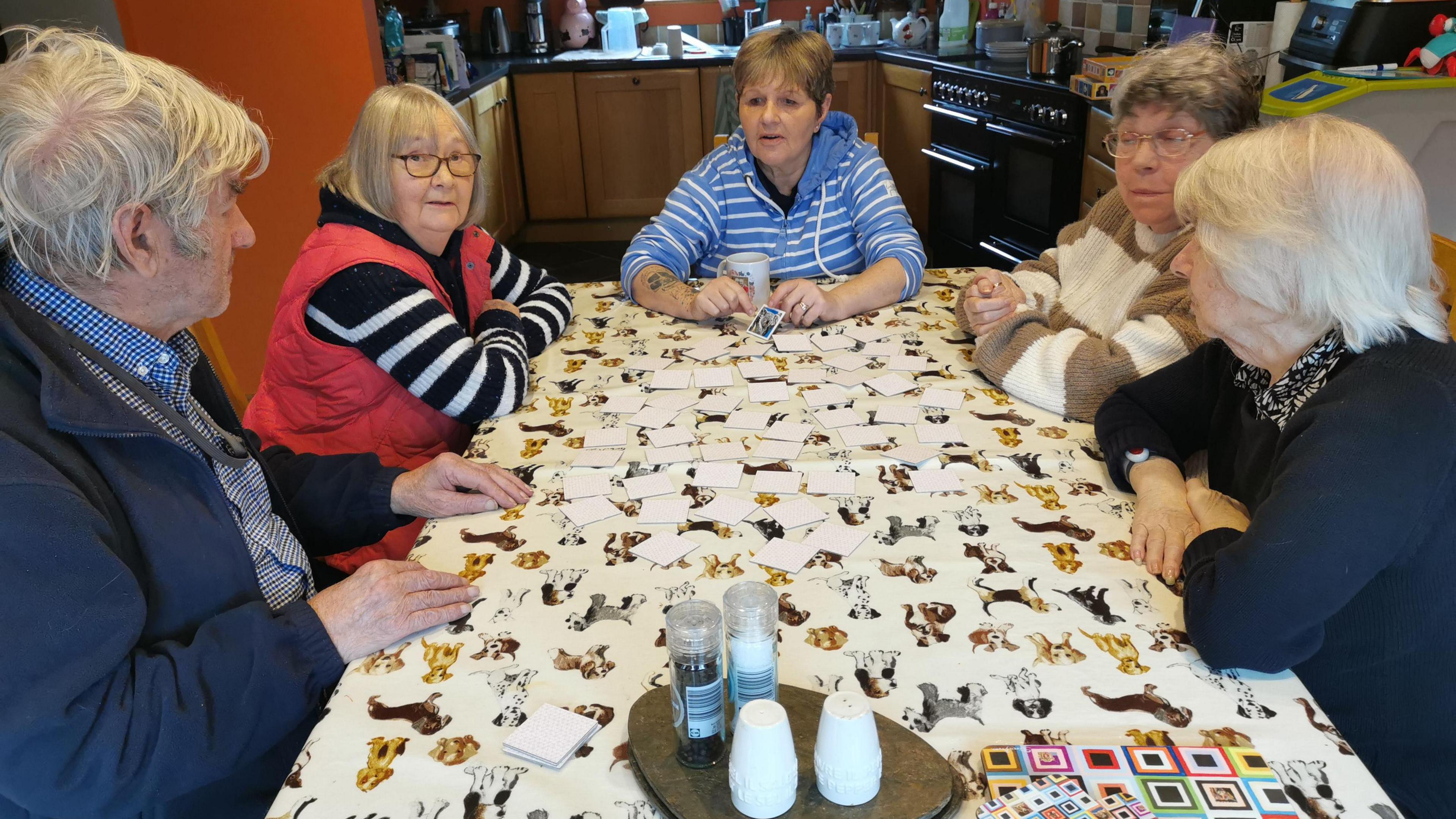 The picture shows host Caroline Acreman sitting at her dining table with a group of four people.  They're taking part in an activity.  In the background is a kitchen.  The tablecloth is covered with pictures of dogs. There are also a salt and pepper pots in the foreground.