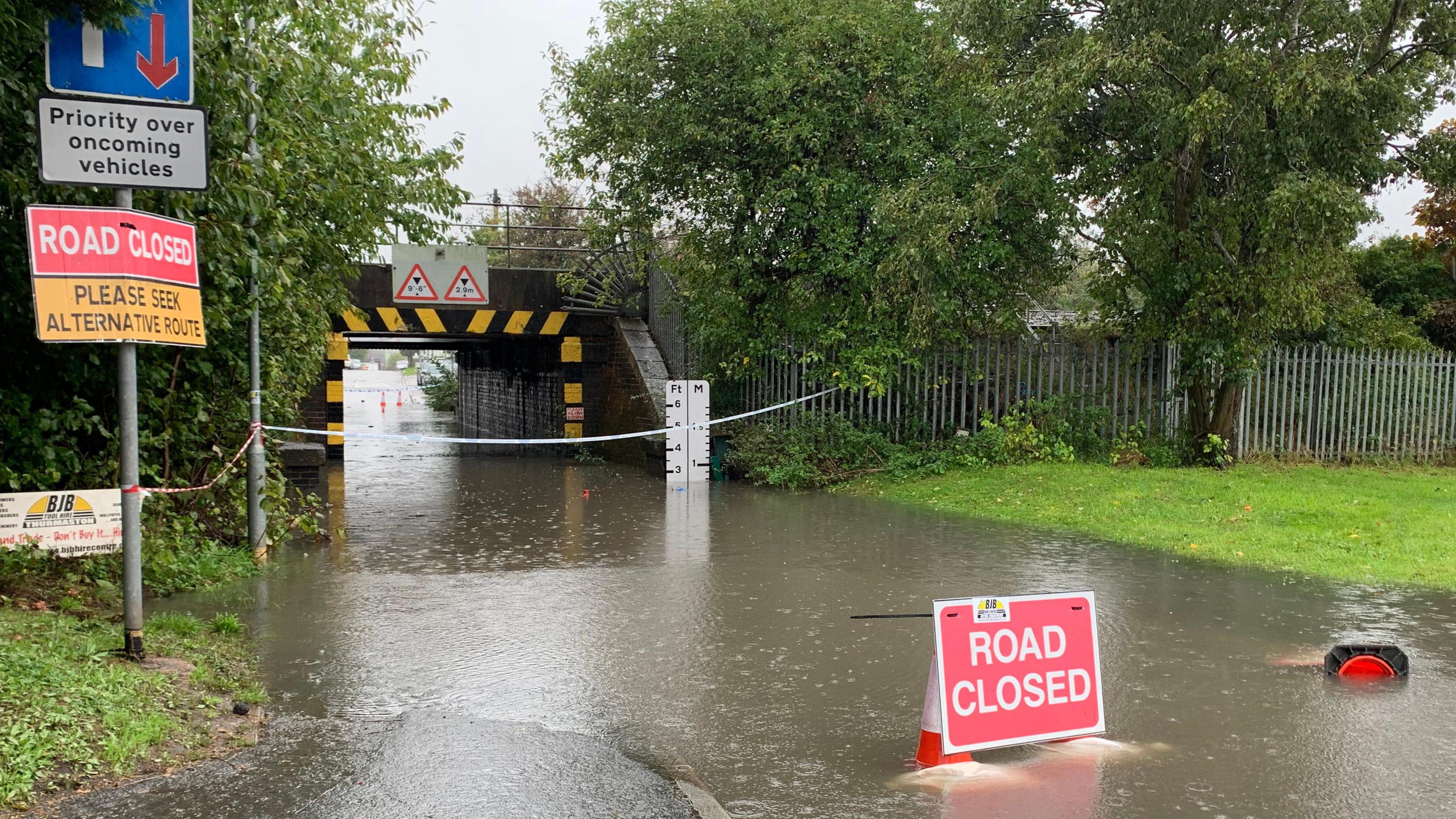 Photograph shows a railway bridge, with flood water beneath it and a flood gauge indicating the depth of the water. There is a road closed sign standing in the water, with an orange overturned cone lying next to the sign