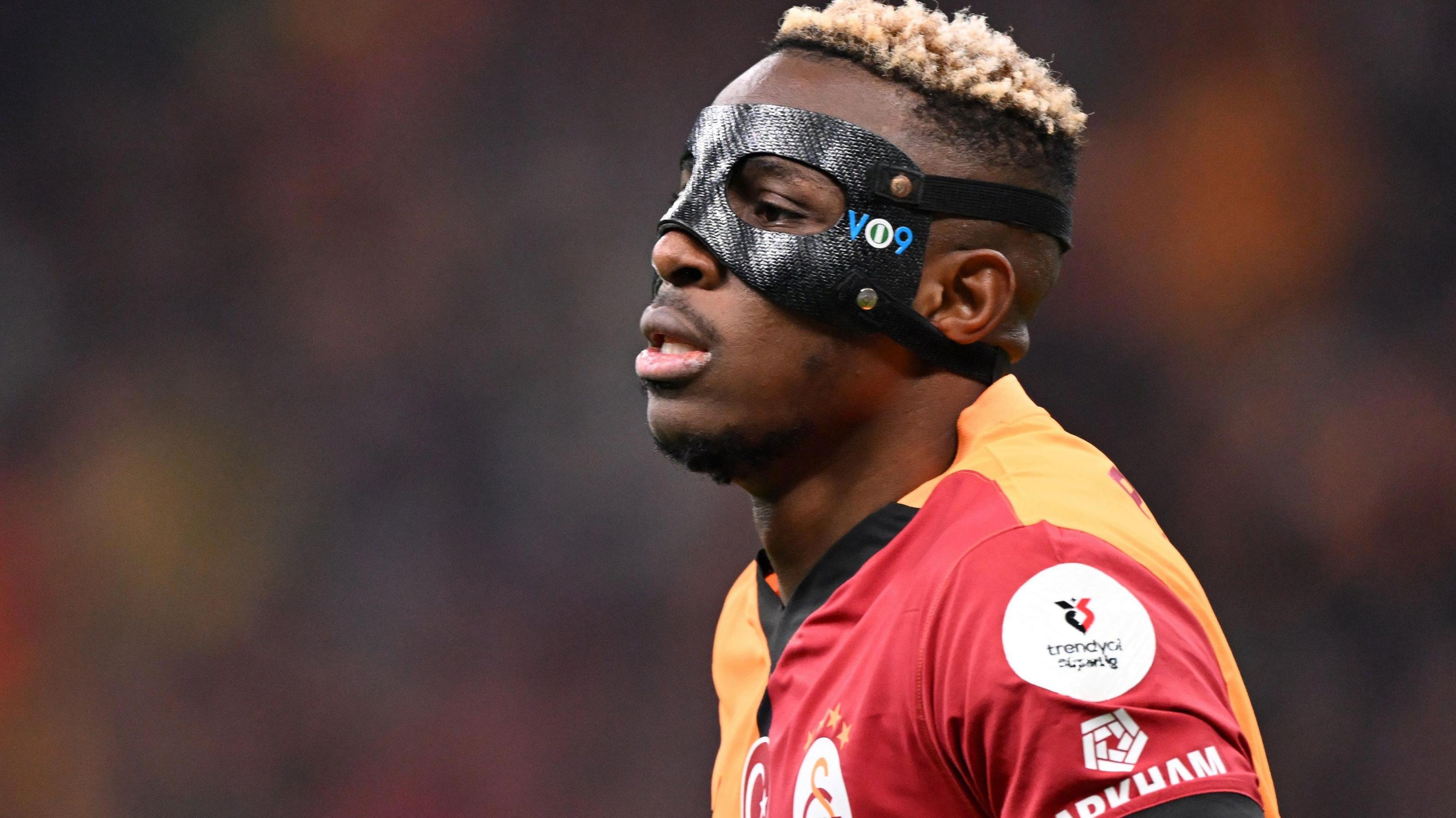 Victor Osimhen wears a red and orange Galatasaray shirt and a protective black eye mask with 'VO9' on the left hand side.