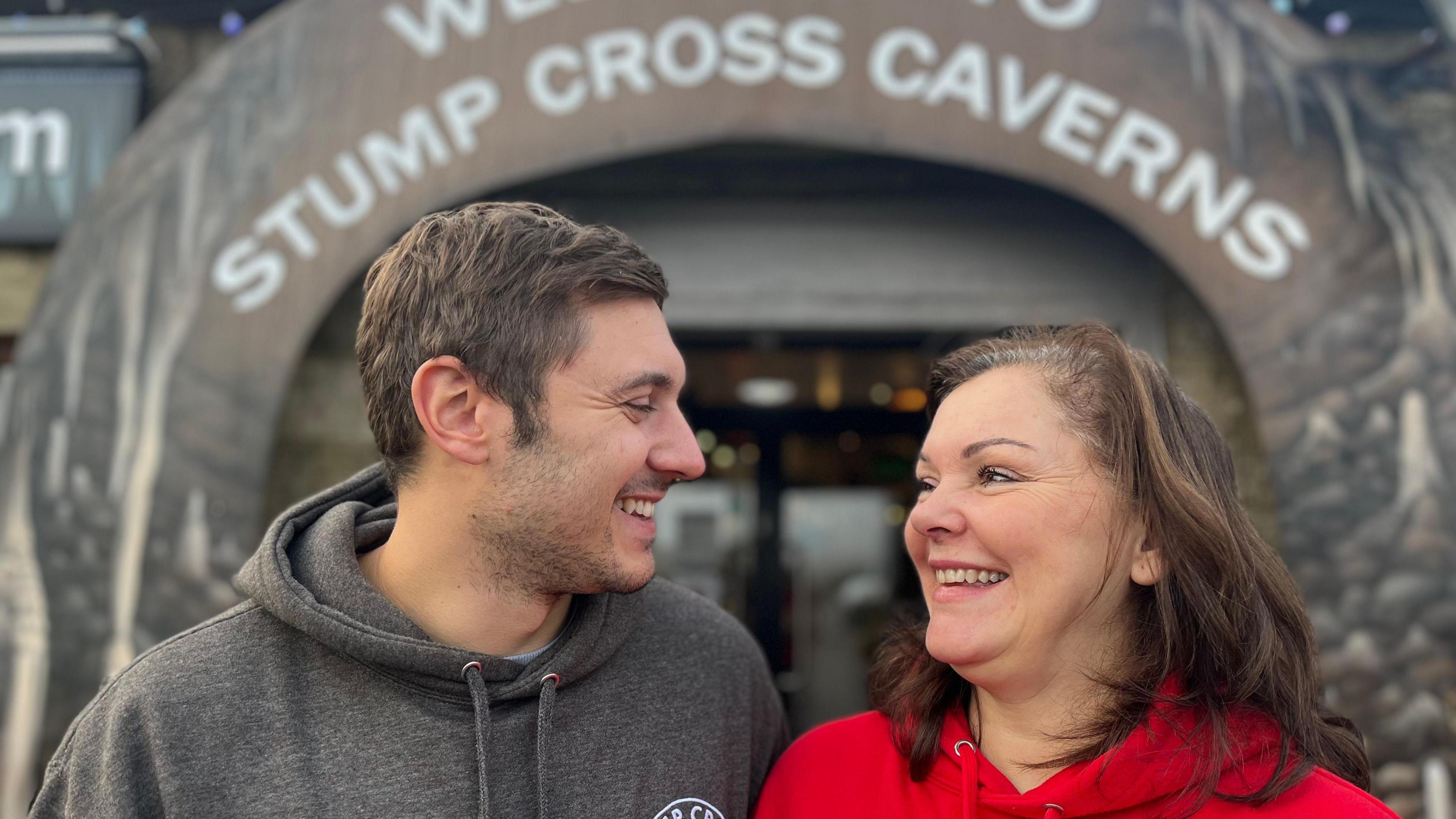 Oliver, who wears a grey hoodie, and his mum Lisa, who wears a red hoodie, look at each other and smile outside a building with a sign reading "Stump Cross Caverns".