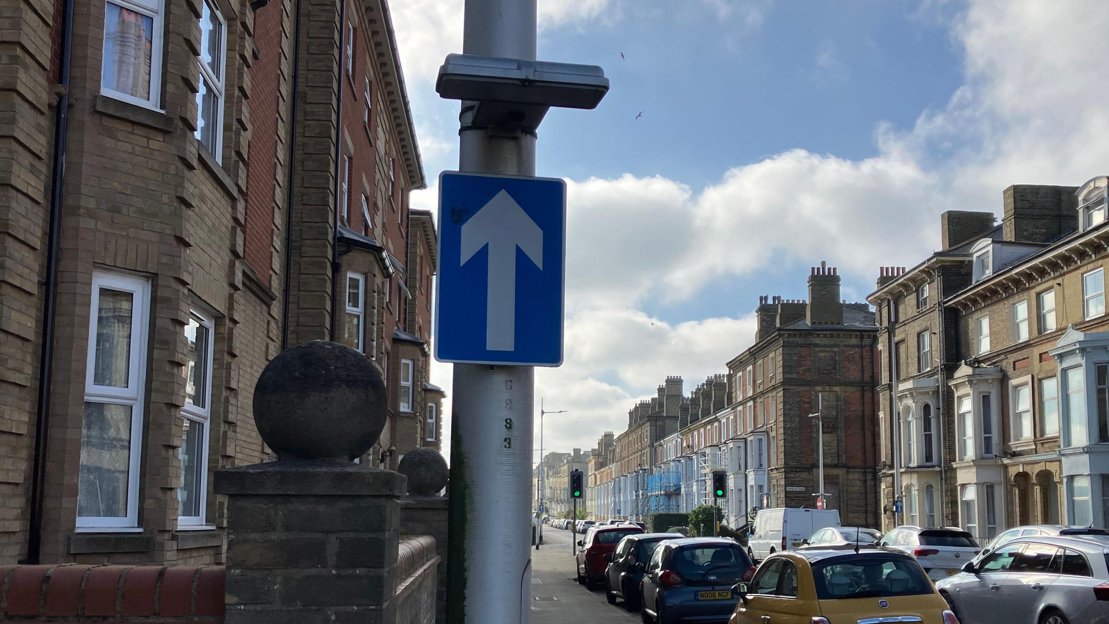 A lamppost in a street with cars parked either side and attached to the lamppost is a blue sign with a white arrow on it indicating it is a one way street