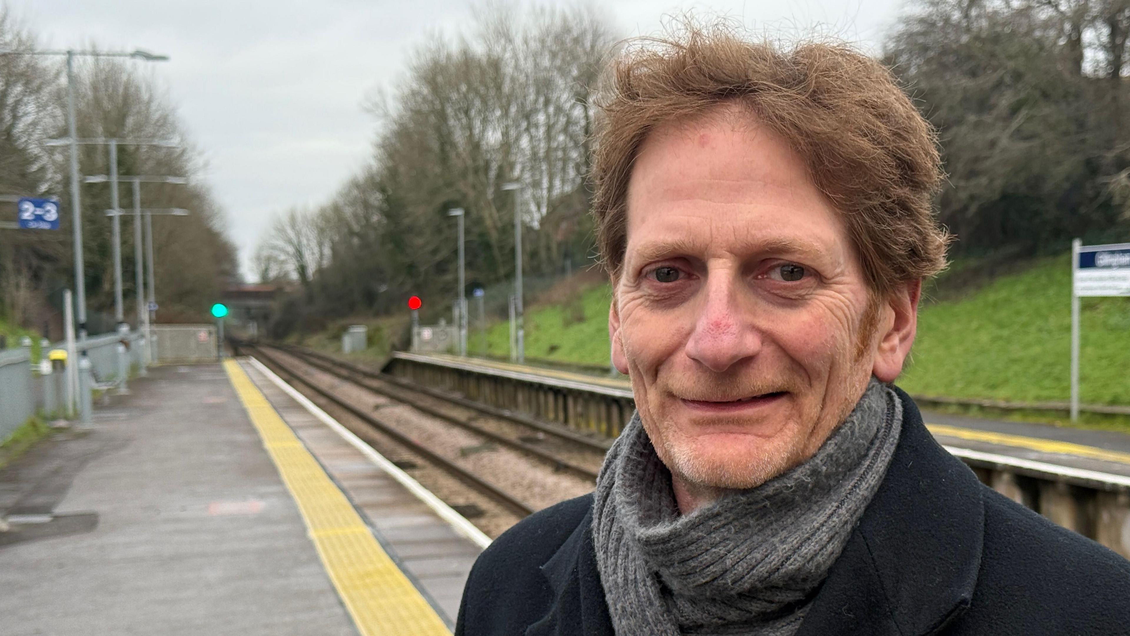 Chris Todd has medium length brown hair, and is wearing a grey scarf and blue overcoat. Behind him out of focus is a train platform and railway line.