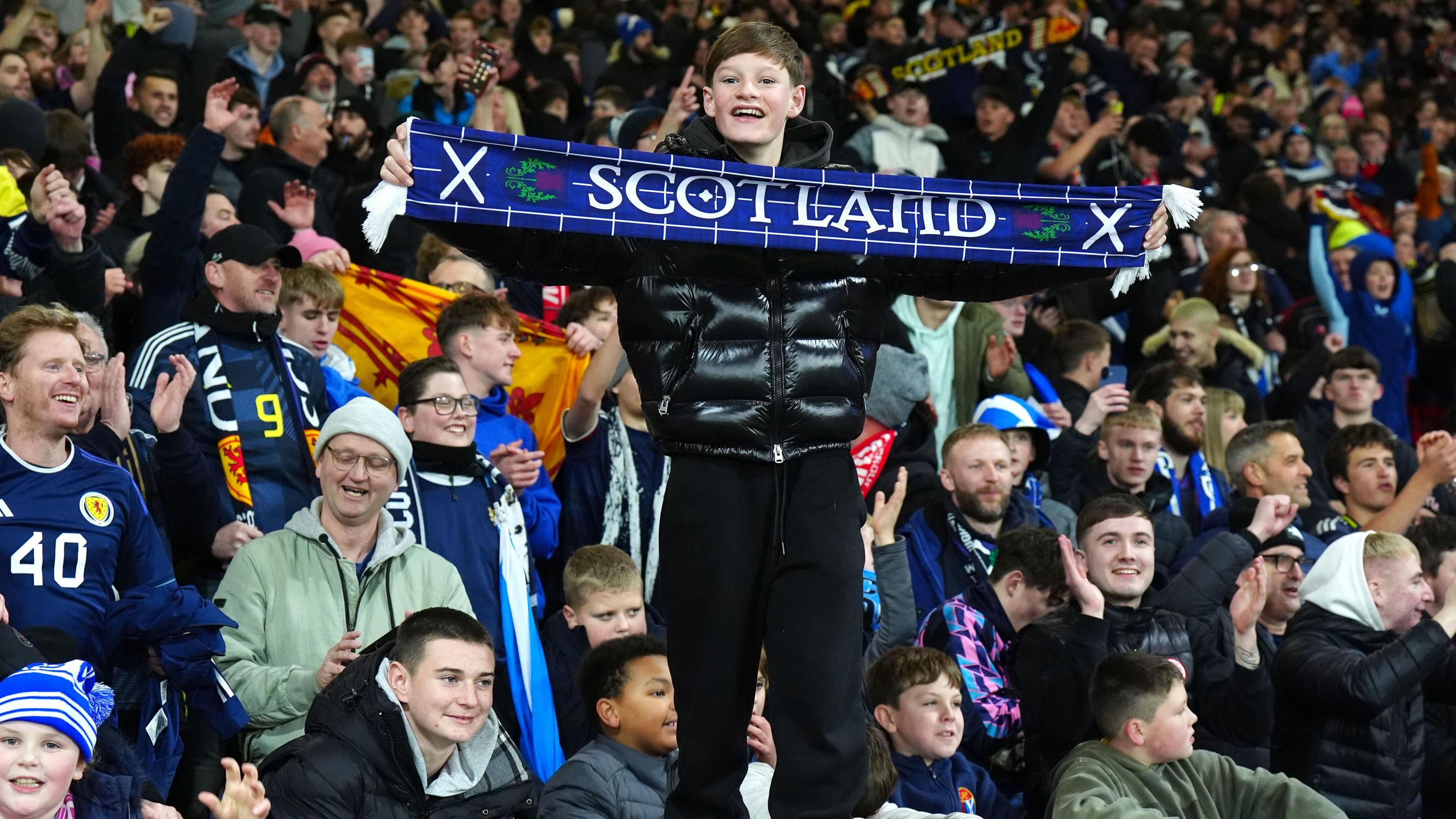 Luchd-leantainn sgioba ball-coise nam fear aig Alba a' dèanamh gàirdeachais agus iad a' faighinn troimhe gu Cupa na Cruinne. Tha fear òg na sheasamh le stoc gorm air a shìneadh aige le Scotland sgrìobhte air 