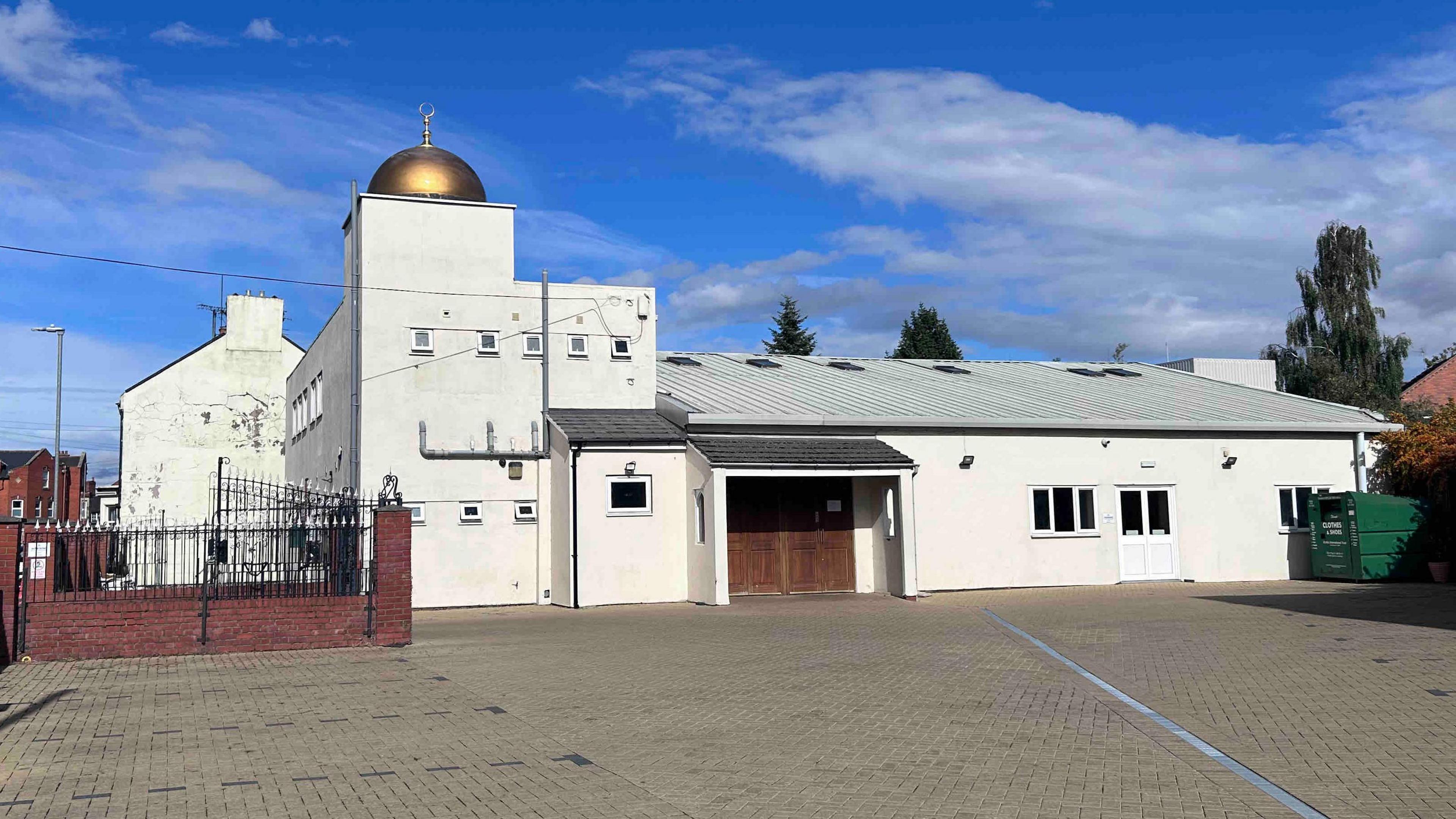 A view of the outside of Northampton Central Mosque on a bright but cloudy day. The mosque is shown from its car park and is mostly white with a gold qubba at its highest point.