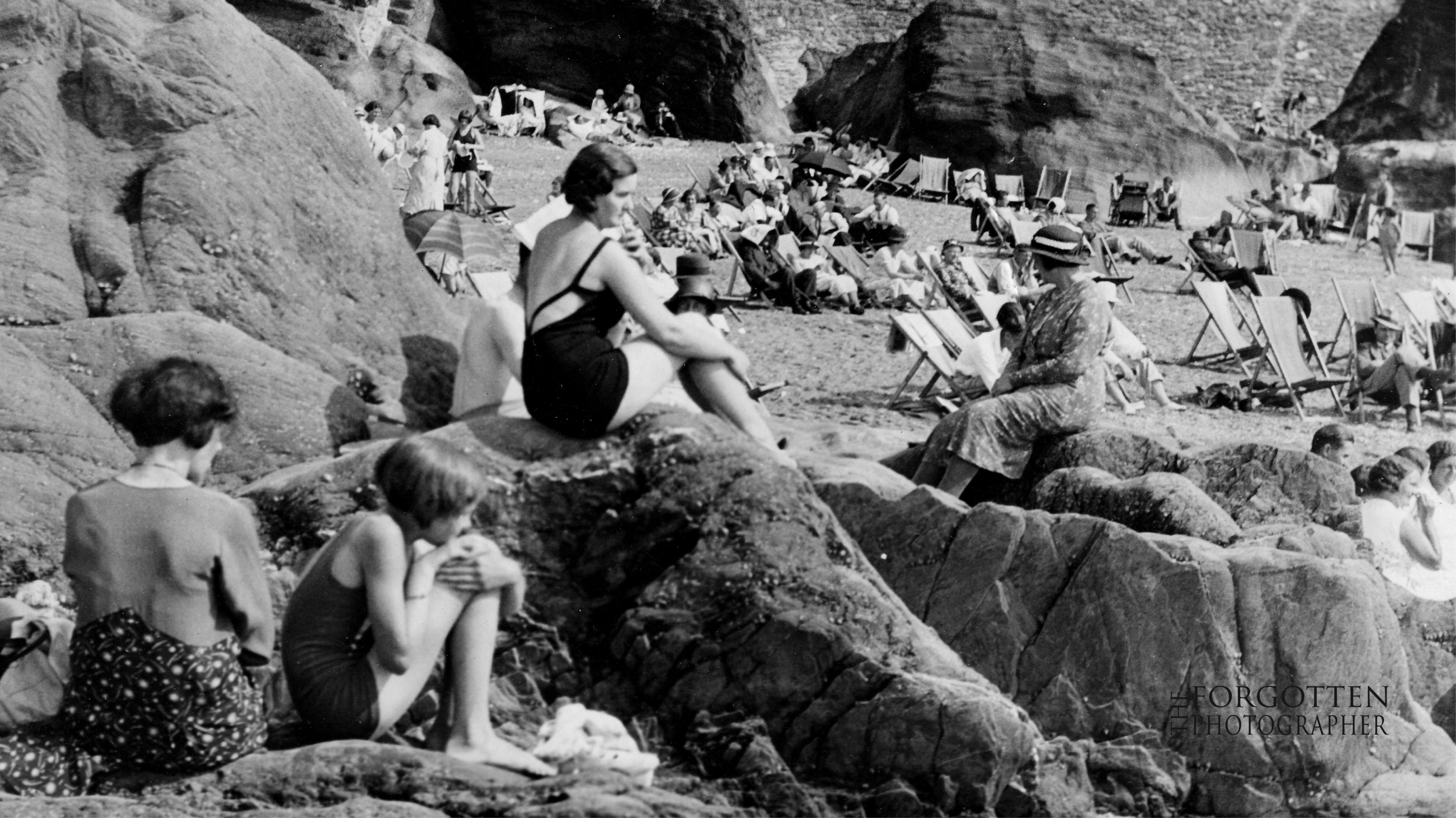 A view of a rocky beach with people lounging on it. Some are wearing swimming costumes and others are fully clothed. They are sitting on decks chairs and rocks. The photo is black and white.