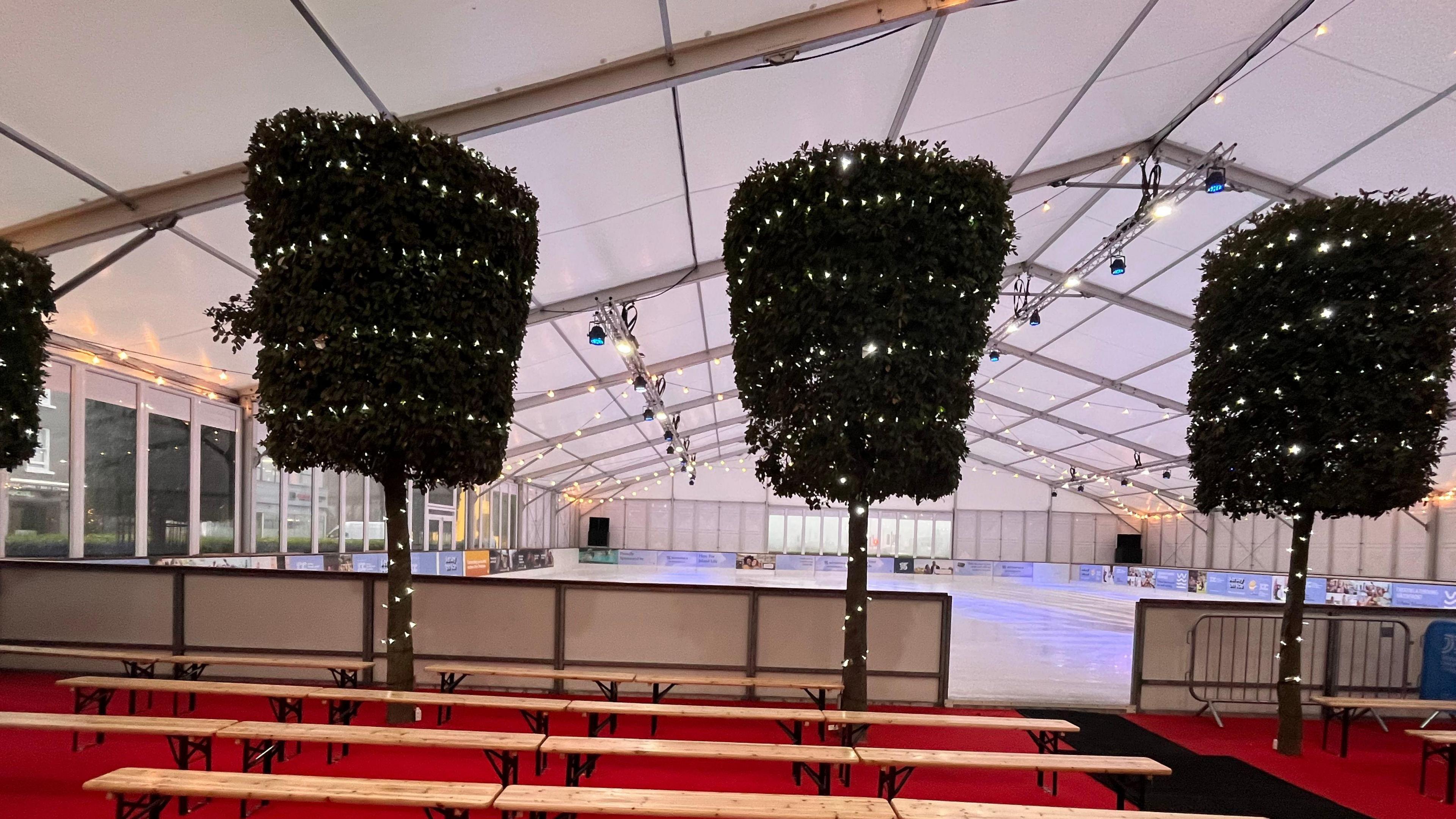 A temporary ice rink inside a marquee, with trees and lights surrounding it.