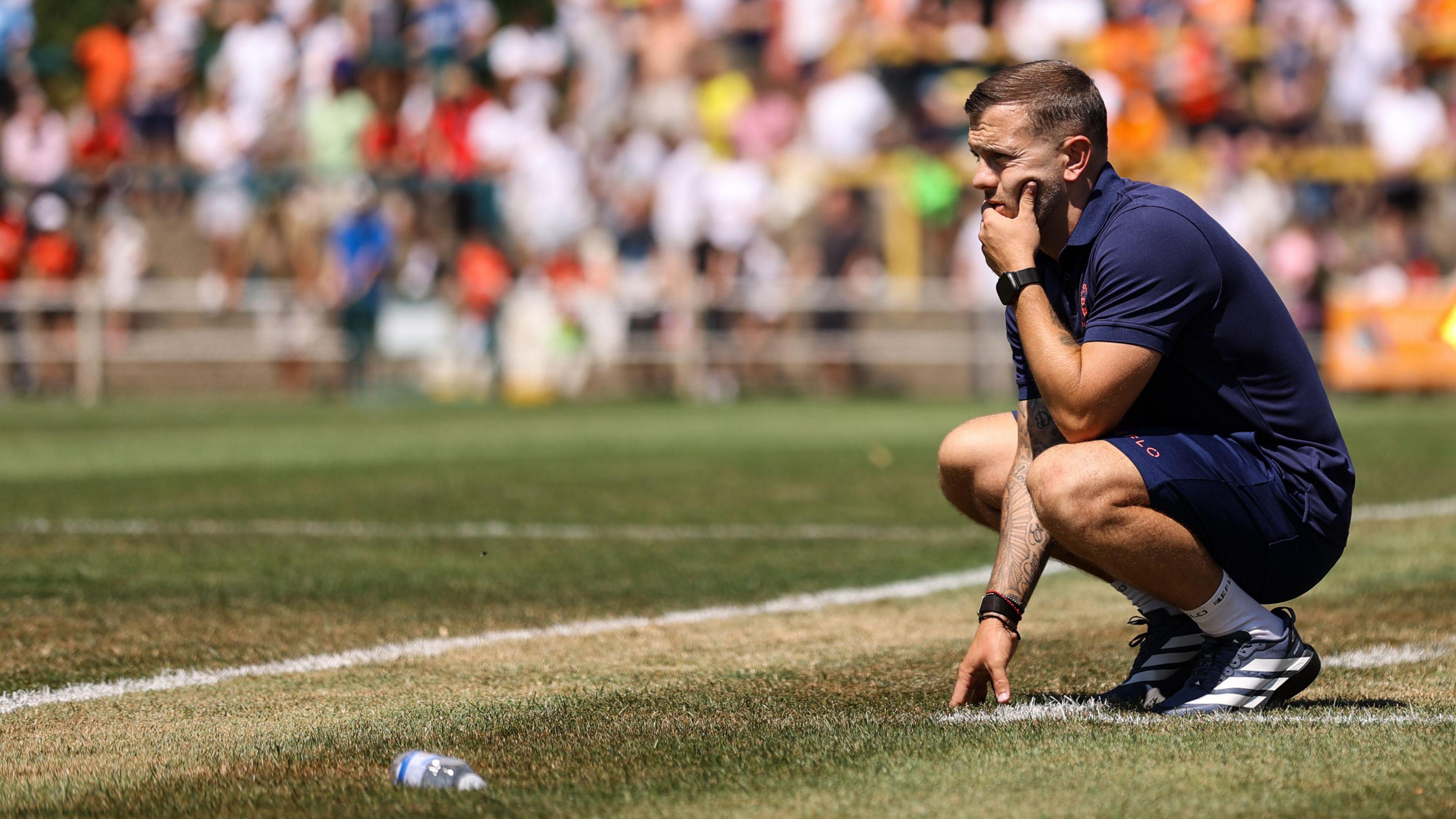 Jack Wilshere, wearing a navy blue outfit and trainers with white socks, crouches down as he watches Luton Town play Hitchin Town in a pre-season friendly in July