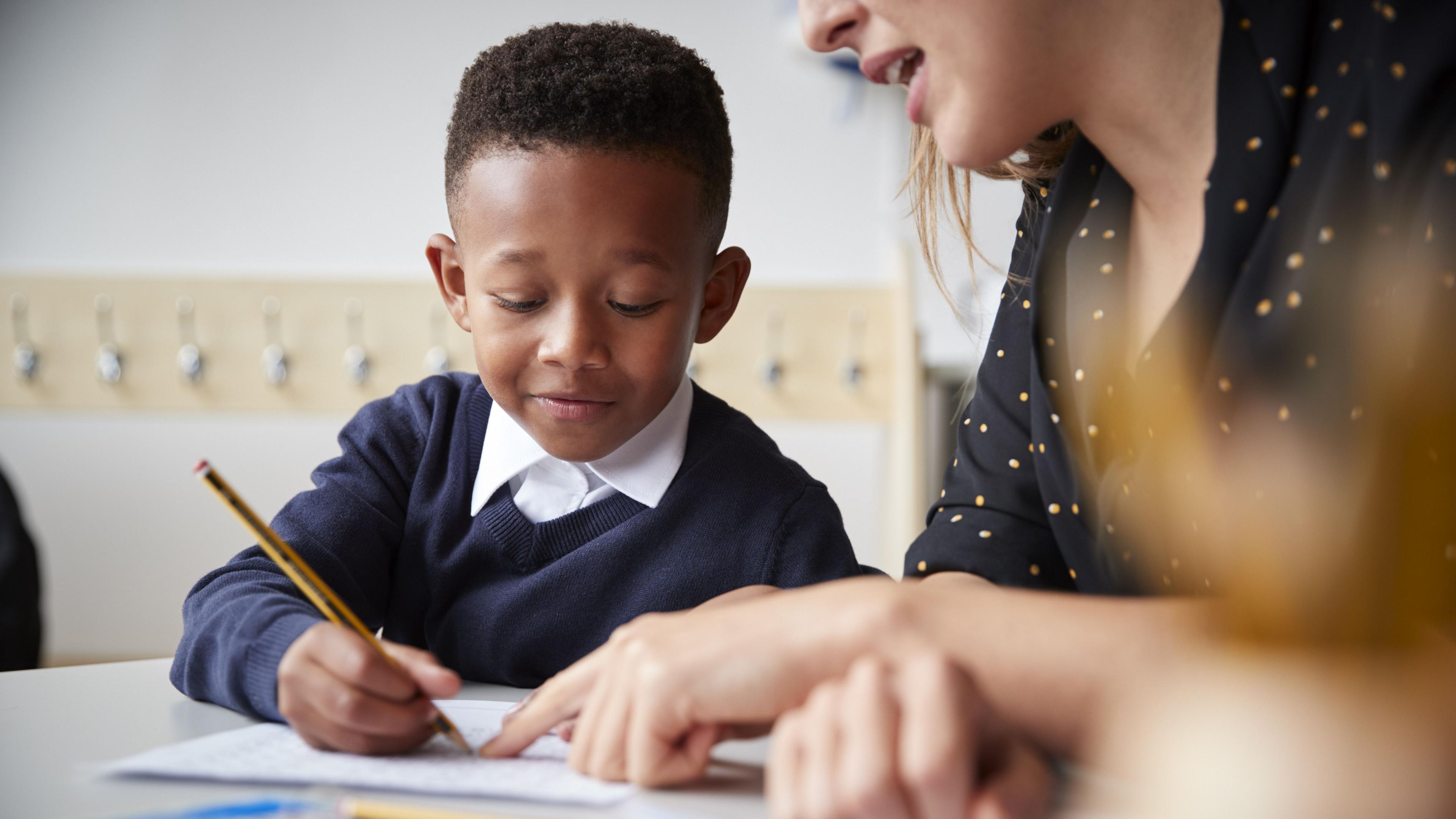 A boy sat at a desk with his teacher, who is helping him with his work as he writes on some paper.