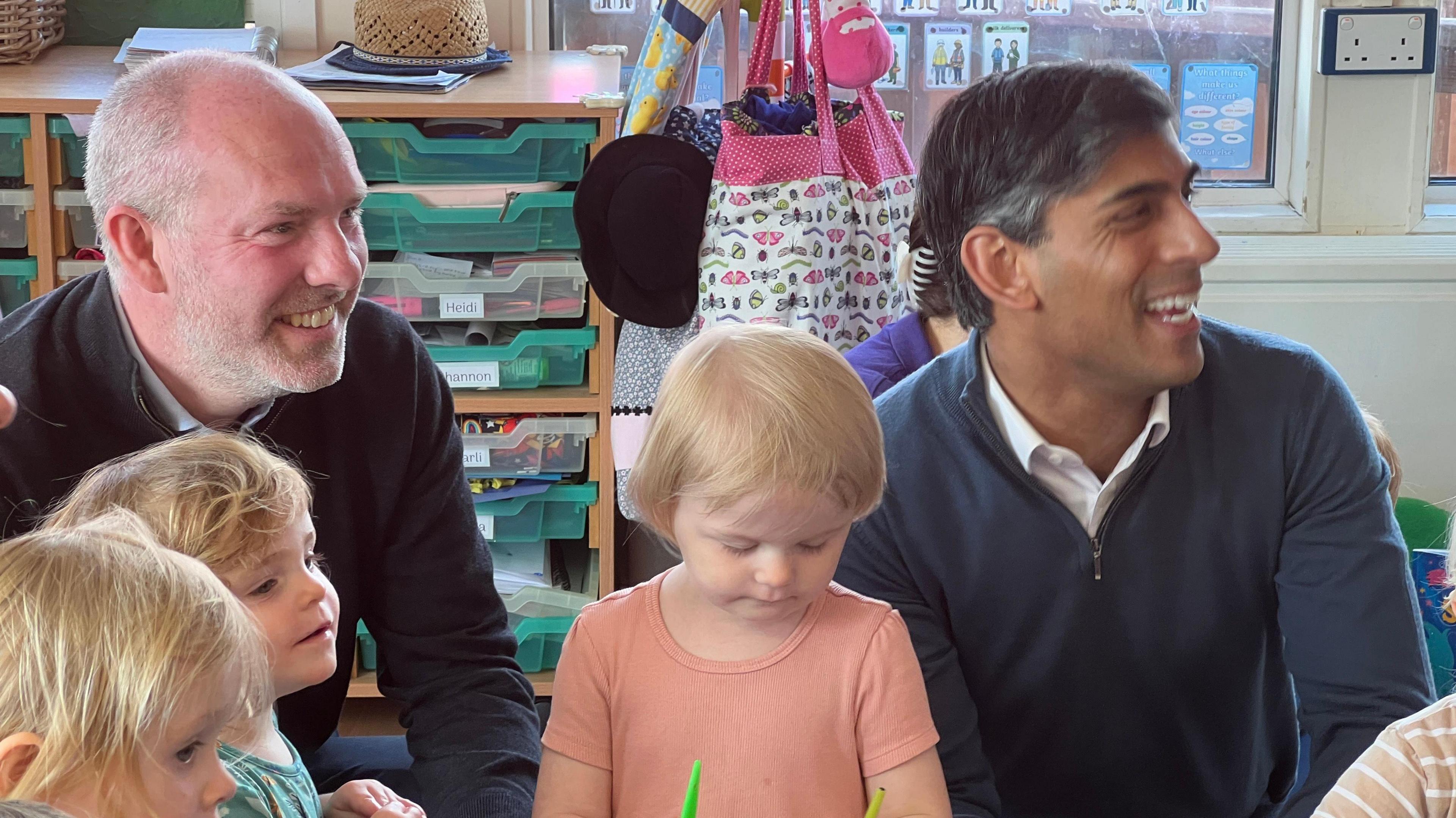 Tomlinson (L) and Rishi Sunak (R) pictured smiling as they talk to nursery children 