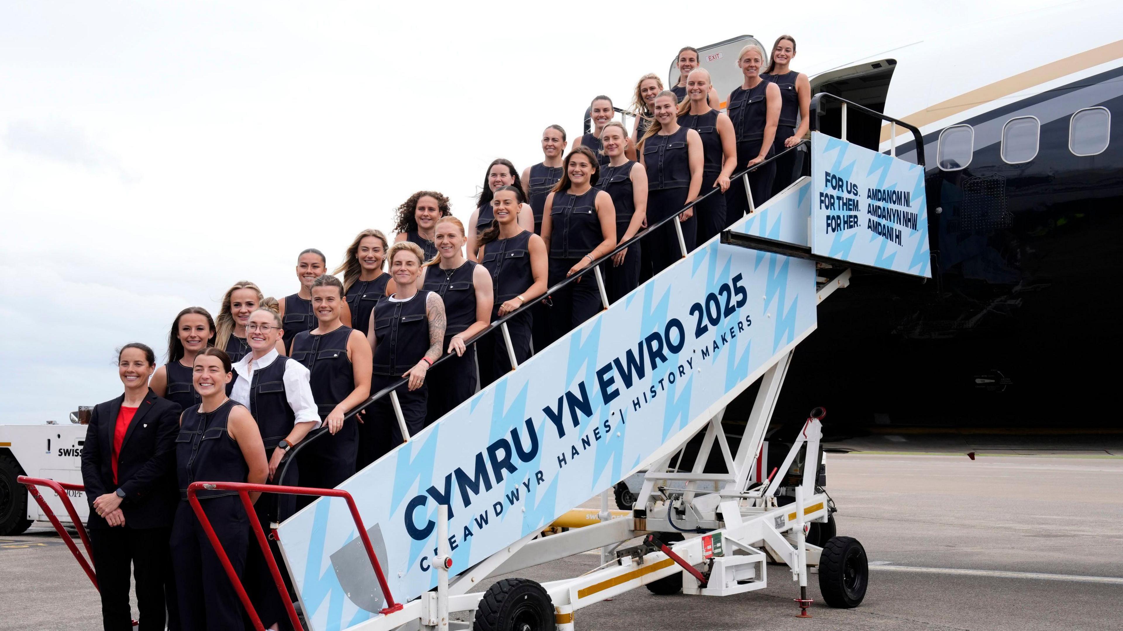 The Wales women's football squad for the 2025 Euros gather on the steps to an airplane. They are all wearing dark sleeveless tops.