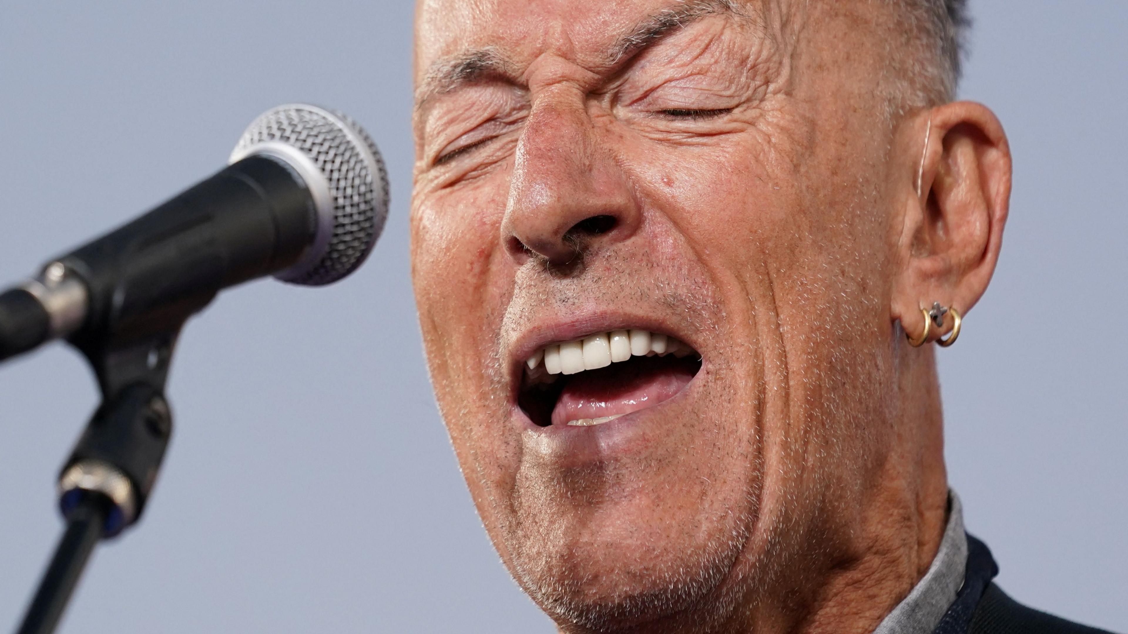 Bruce Sprungsteen sings into a microphone. He has grey hair and very thin grey stubble, with a gold earring in his left ear
