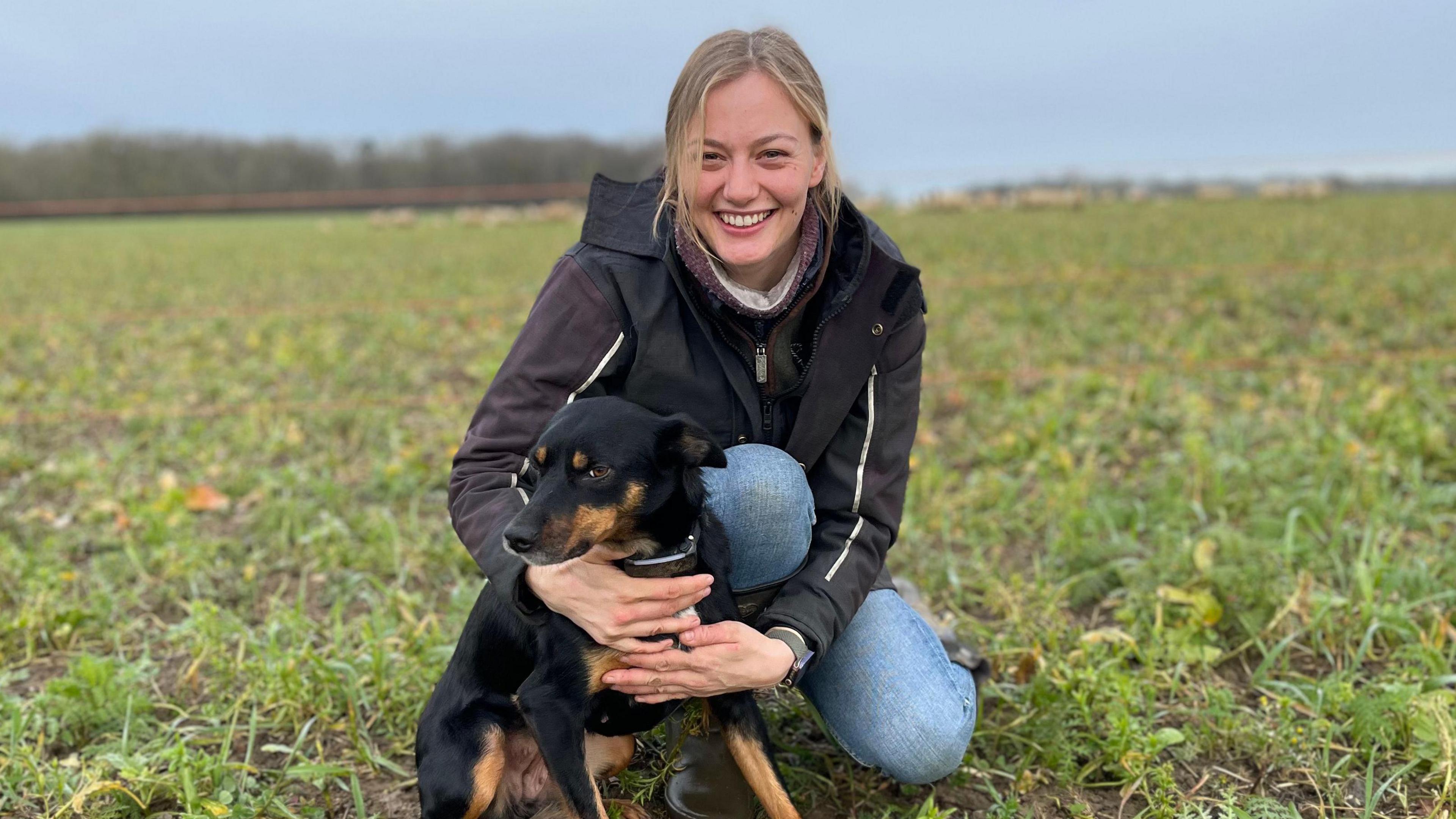 Hannah Murrell a woman with blonde hair is crouched down in a field with her black and brown sheepdog Roo. She is wearing blue jeans and a black coat. She is looking at the camera and is smiling. 