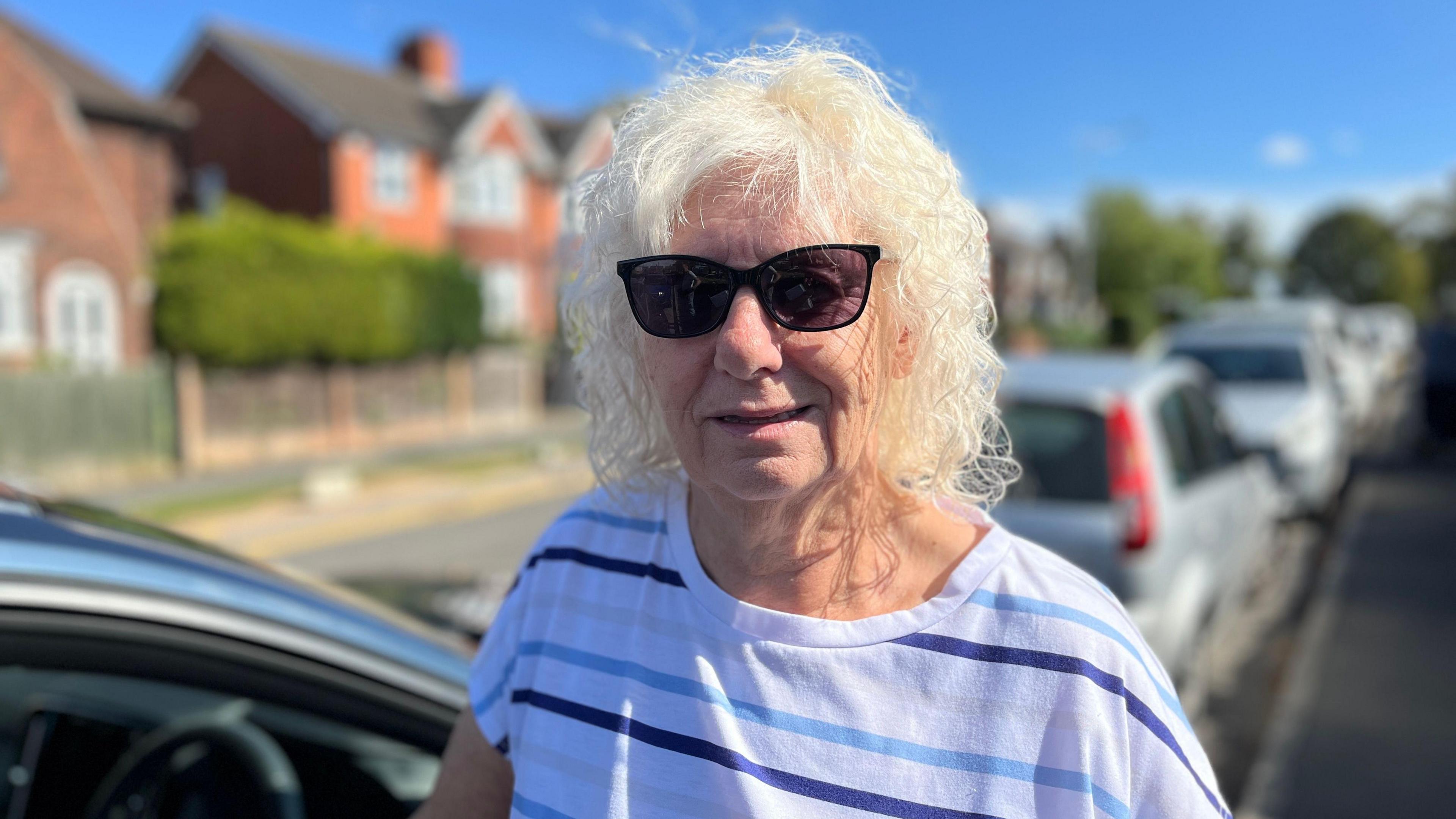 A woman with white curly short hair and black sunglasses is wearing a stripey white and blue top and looking at the camera next to a car. The background is blurred and there is a road with houses in sight.