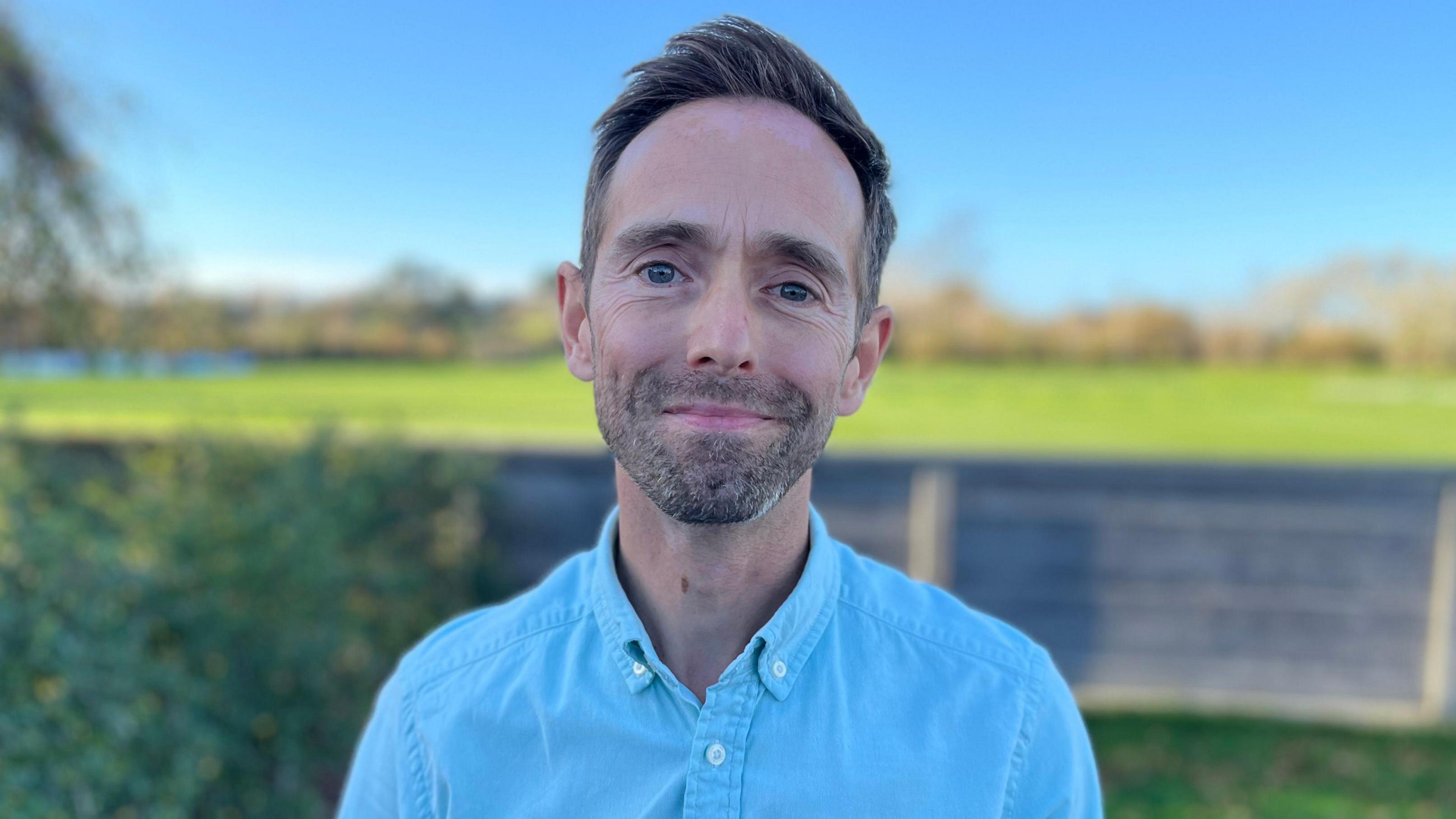 Dr Simon Sebire - A man with coifed brown haird and a stubbly beard with a fleck of grey on the bottom. He is smiling at the camera and has a blue buttoned shirt on.