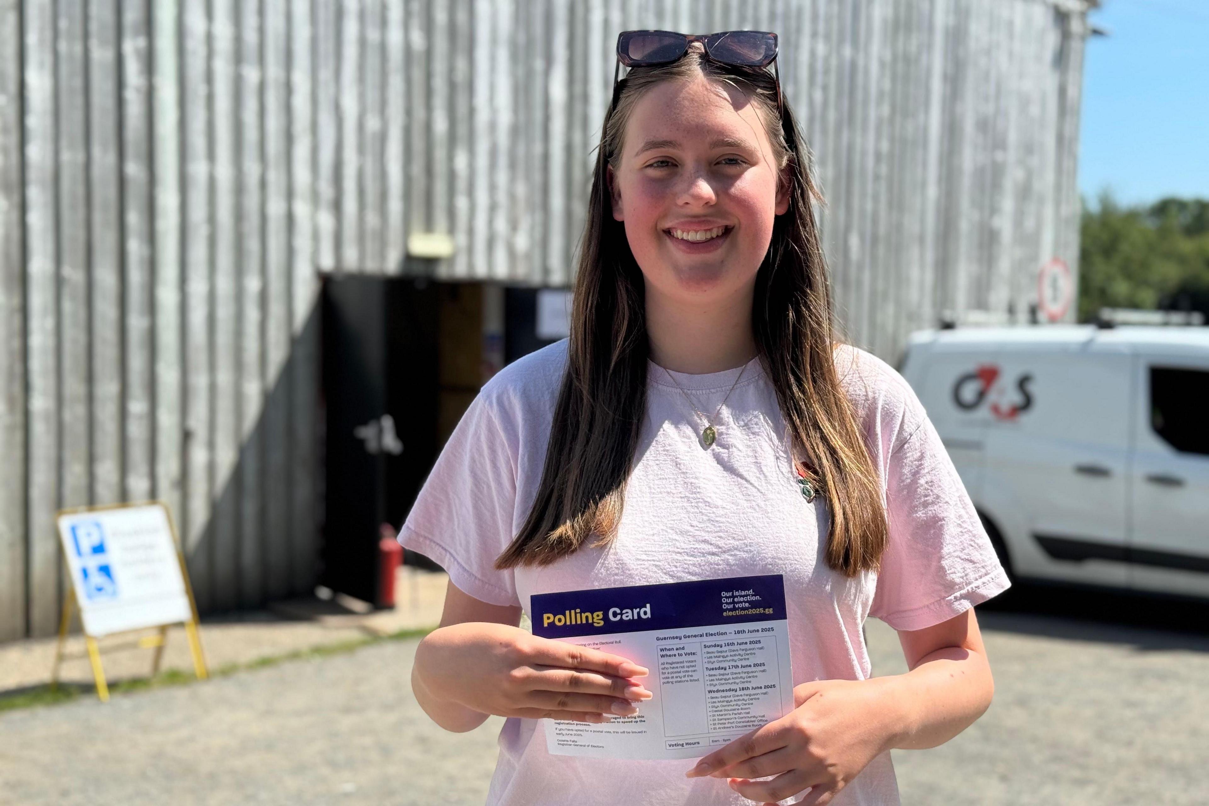 A young women with long brown hair smiling at the camera. She is wearing a pale pink top and has sunglasses resting on her head. She is hold her polling card up to the camera.