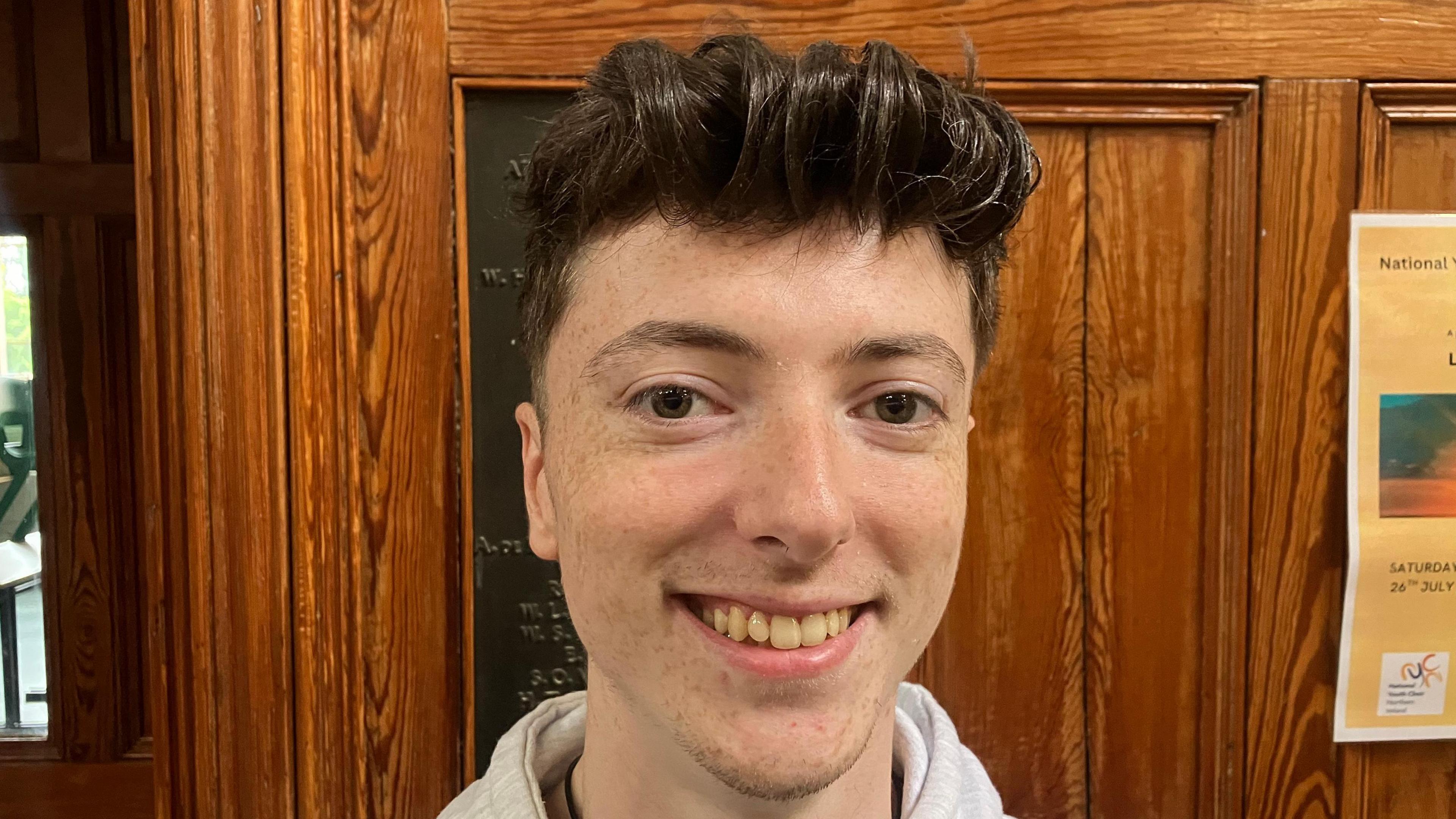 Daniel Stewart is smiling at the camera in front of a wooden door panel. He has short dark hair, brown eyes and is wearing a grey hoodie. 