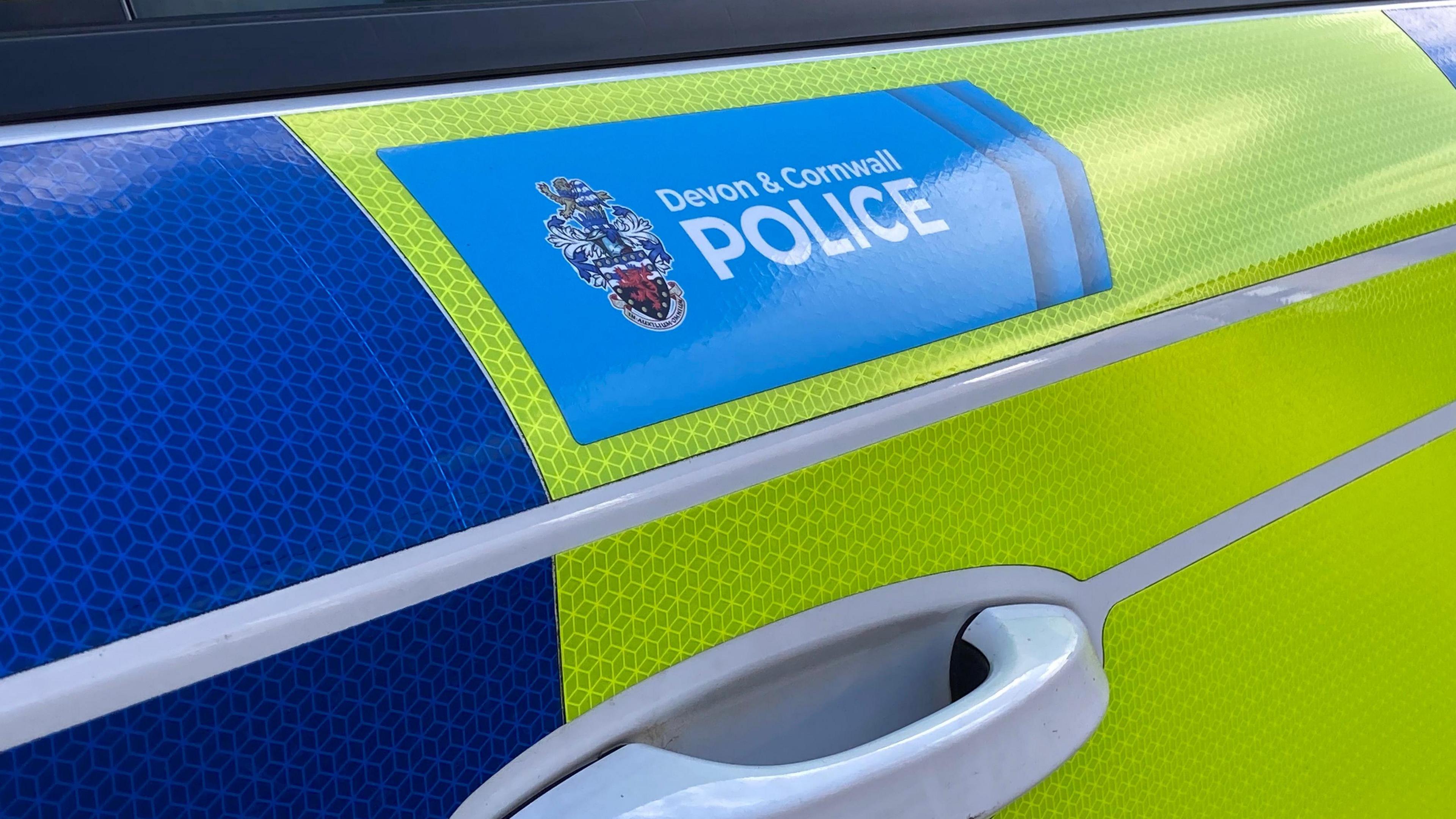 The side of a blue and yellow police vehicle. Above the white door handle there is a blue box with the words 'Devon and Cornwall Police' with a badge beside it.