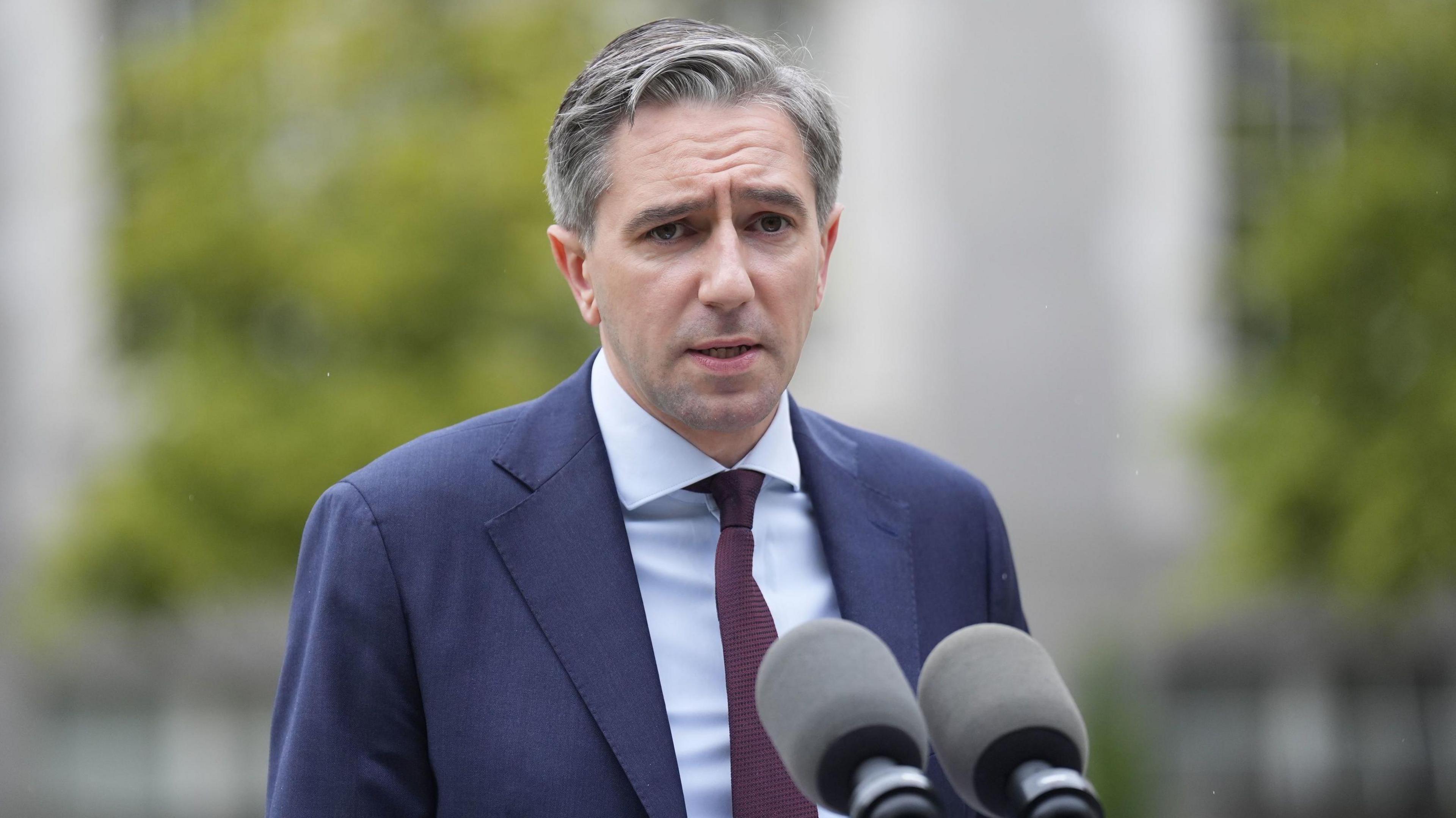 Simon Harris stands at a podium with two microphones placed in front of him. He has short grey hair, and is wearing a navy suit jacket, a light blue buttoned shirt and dark purple tie.