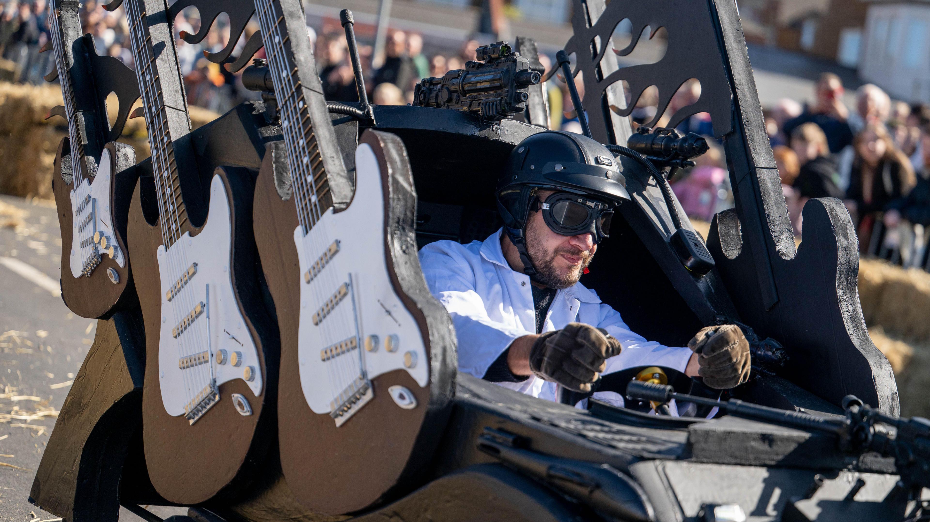 A kart which has been designed with large black and white detailed guitars on the side of it. The driver inside the kart is wearing a white jacket, brown gloves, a black helmet and eye goggles.