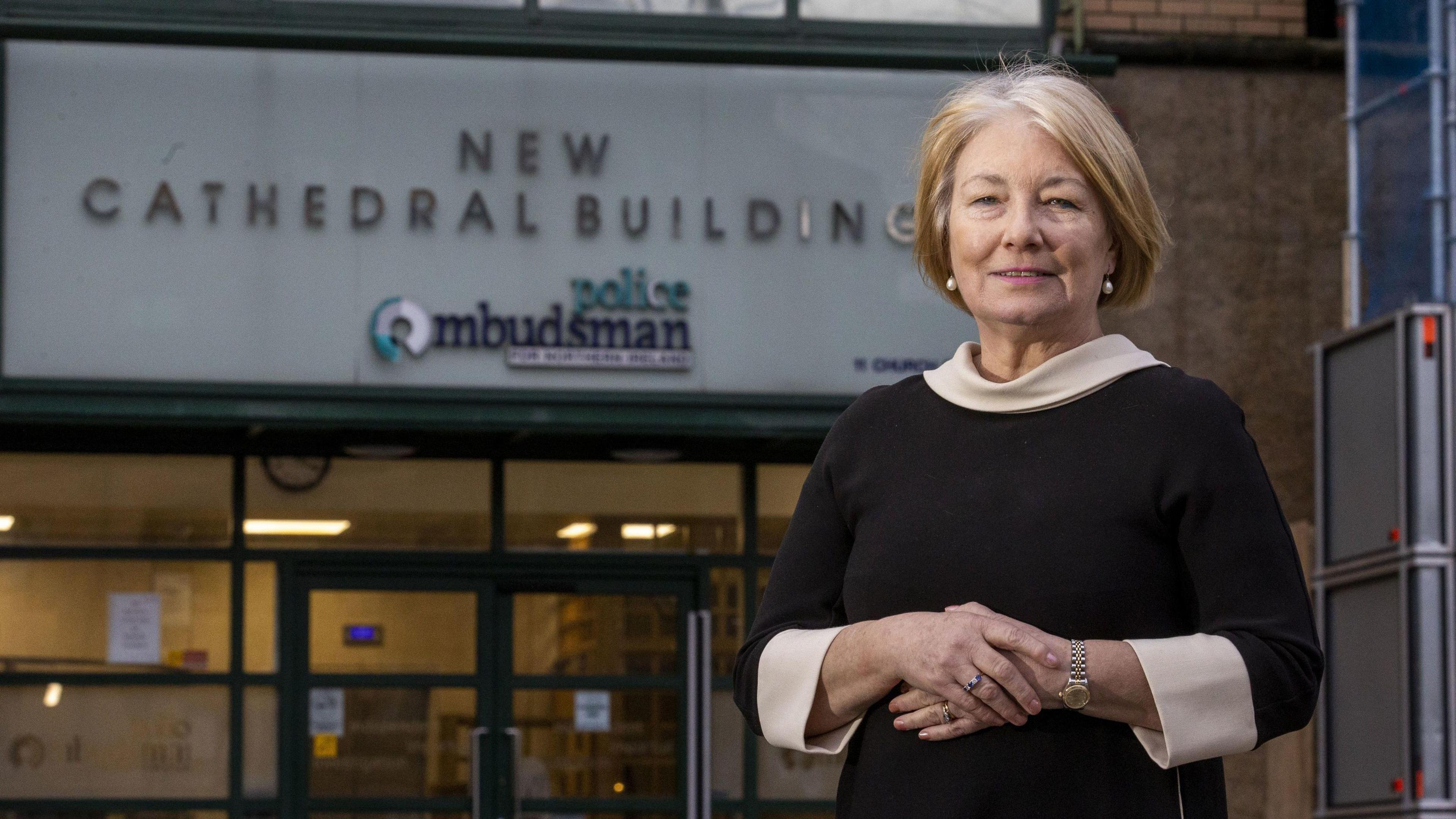 Marie Anderson standing outside the police ombudsman office in Belfast. She wears a black dress with a white collar and cuffs. She has short, blonde hair.