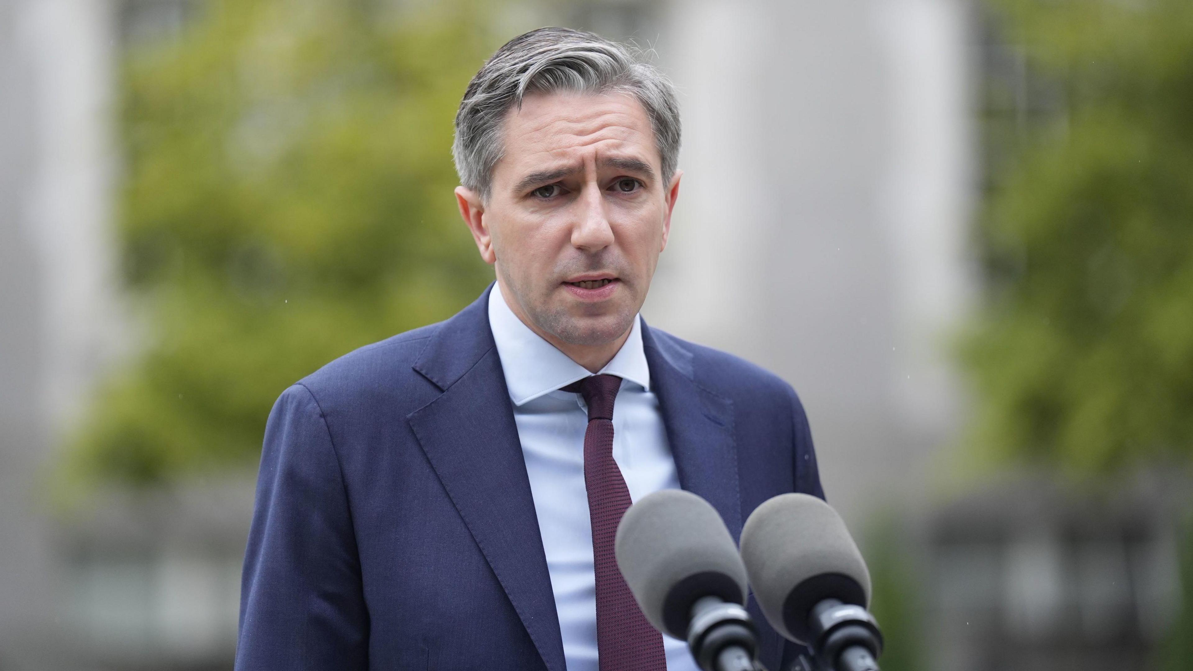 Simon Harris wearing a blue shirt, burgundy tie and navy suit. He is standing in front of a microphone.