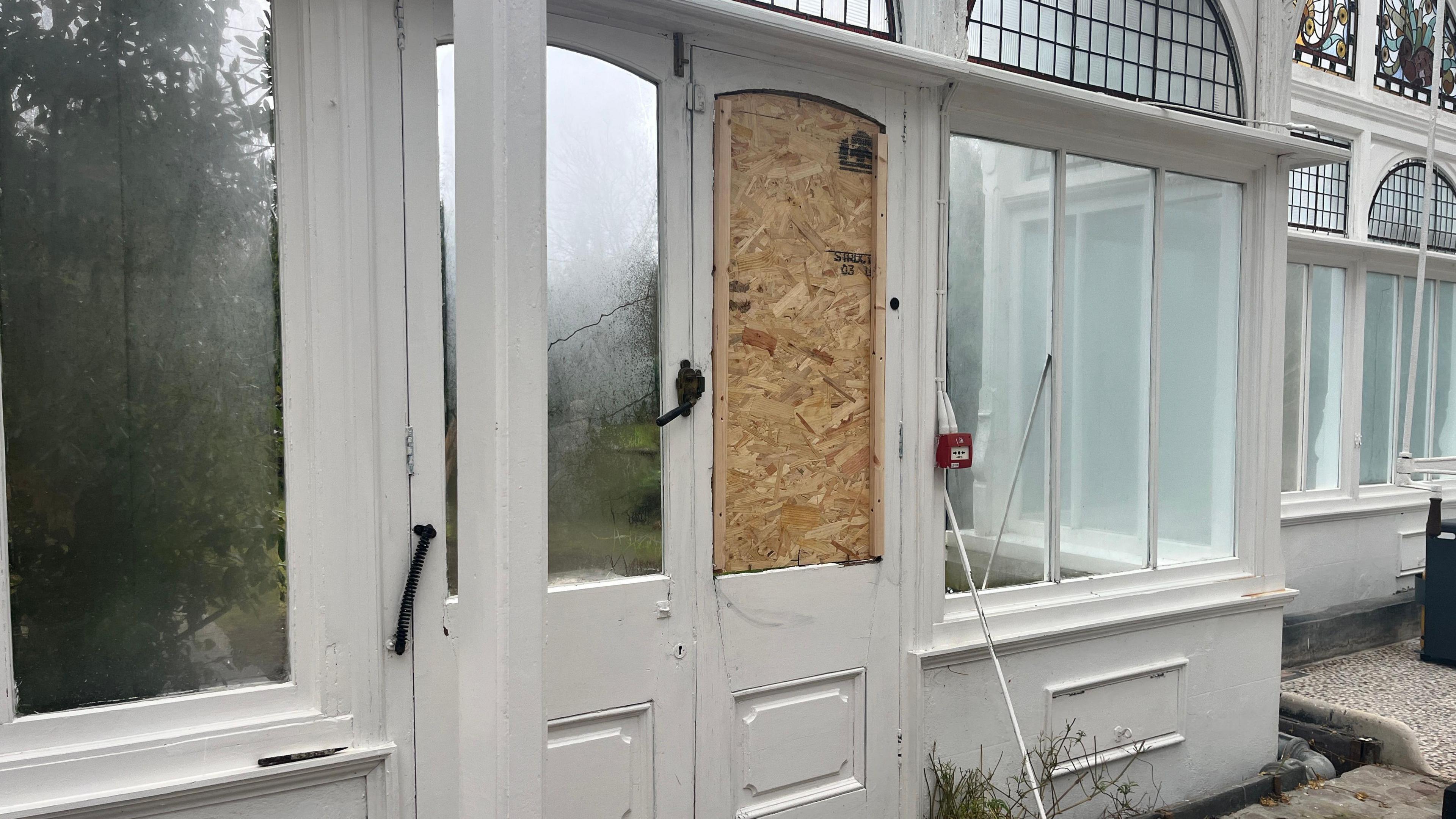 The doors inside a light conservatory. The wall is lined with windows, and one set of double doors. One of the doors has it's window blocked with a wooden board. Below the windows, the wall is white and there is a white tiled floor. 