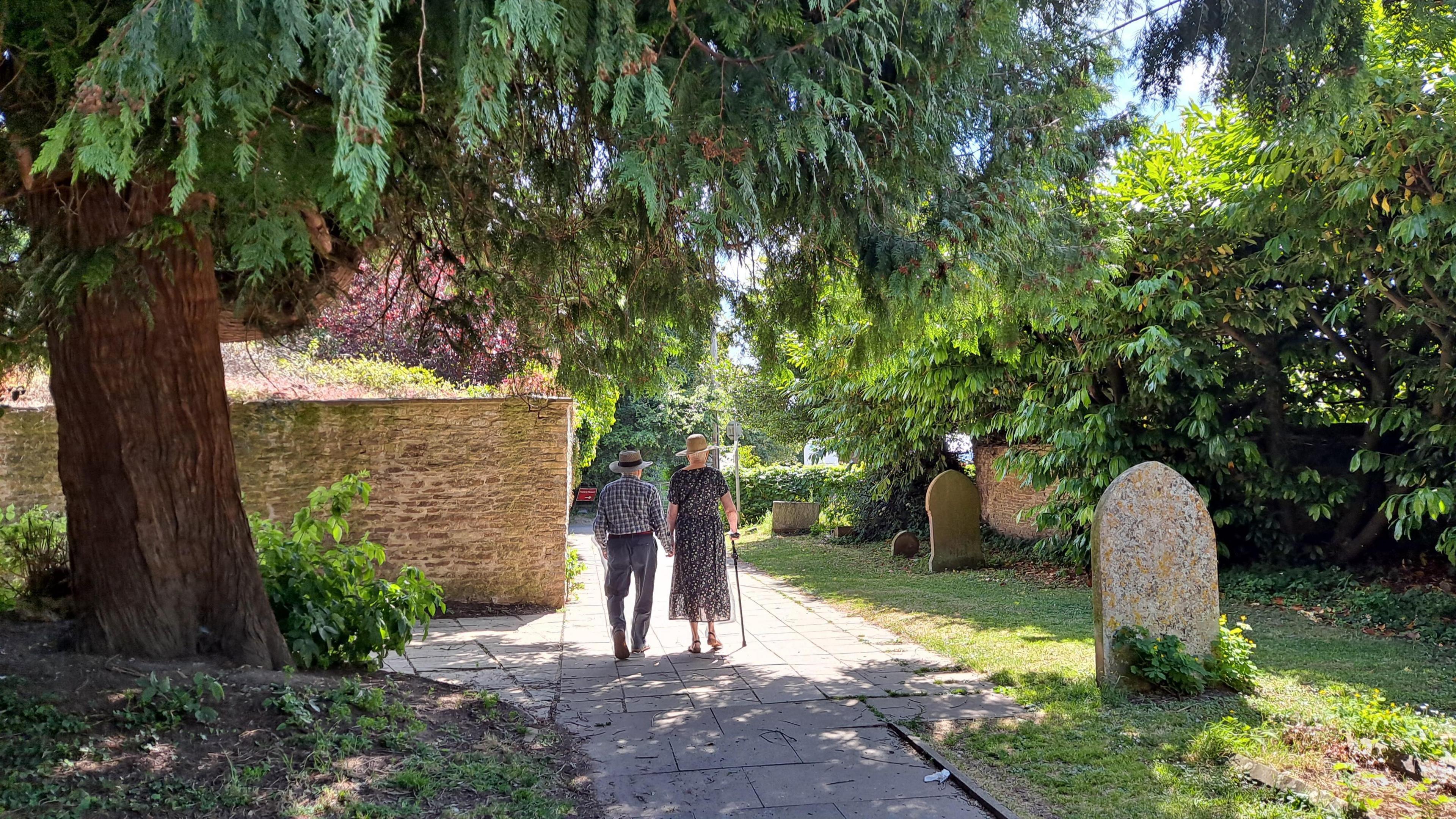 WEDNESDAY - A man and woman hold hands walk through a graveyard on a sunny day. They are both wearing straw hats. The woman is using a walking stick and is wearing a dress, the man is wearing a shirt and trousers. They are walking under a pine tree into the sunshine. On their left is a brick wall, on the right are several gravestones and a tall green hedge.