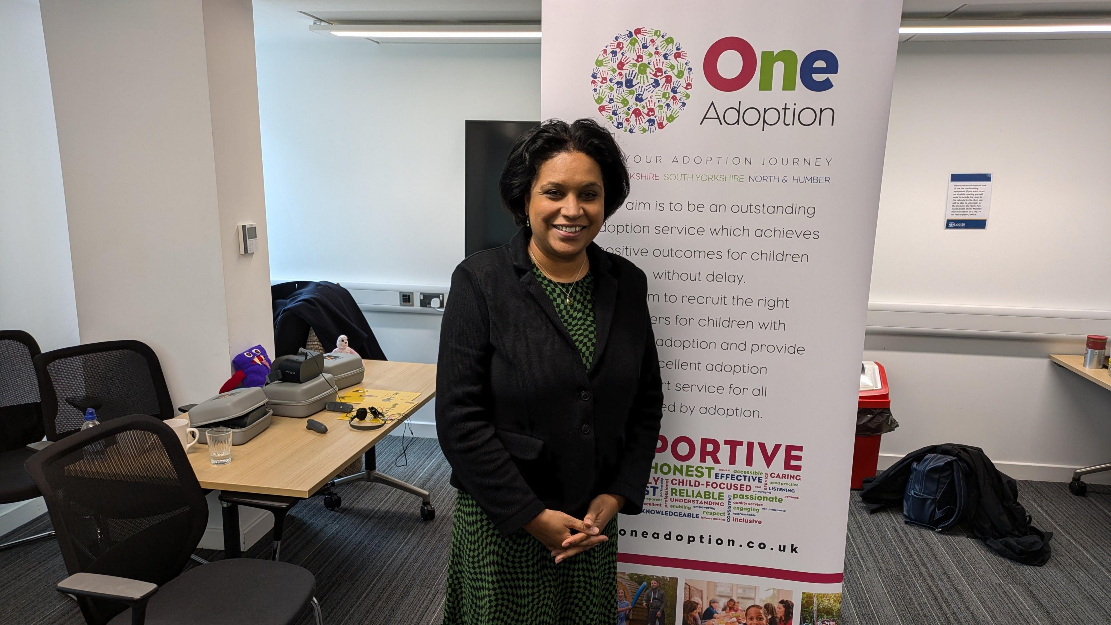 Children's minister Janet Daby, a lady with dark hair, a black jacket and a green and black dress. She is stood partially in front of an advertising board for One Adoption, which has large writing on explaining what the agency is. A desk with various items is over her right shoulder.