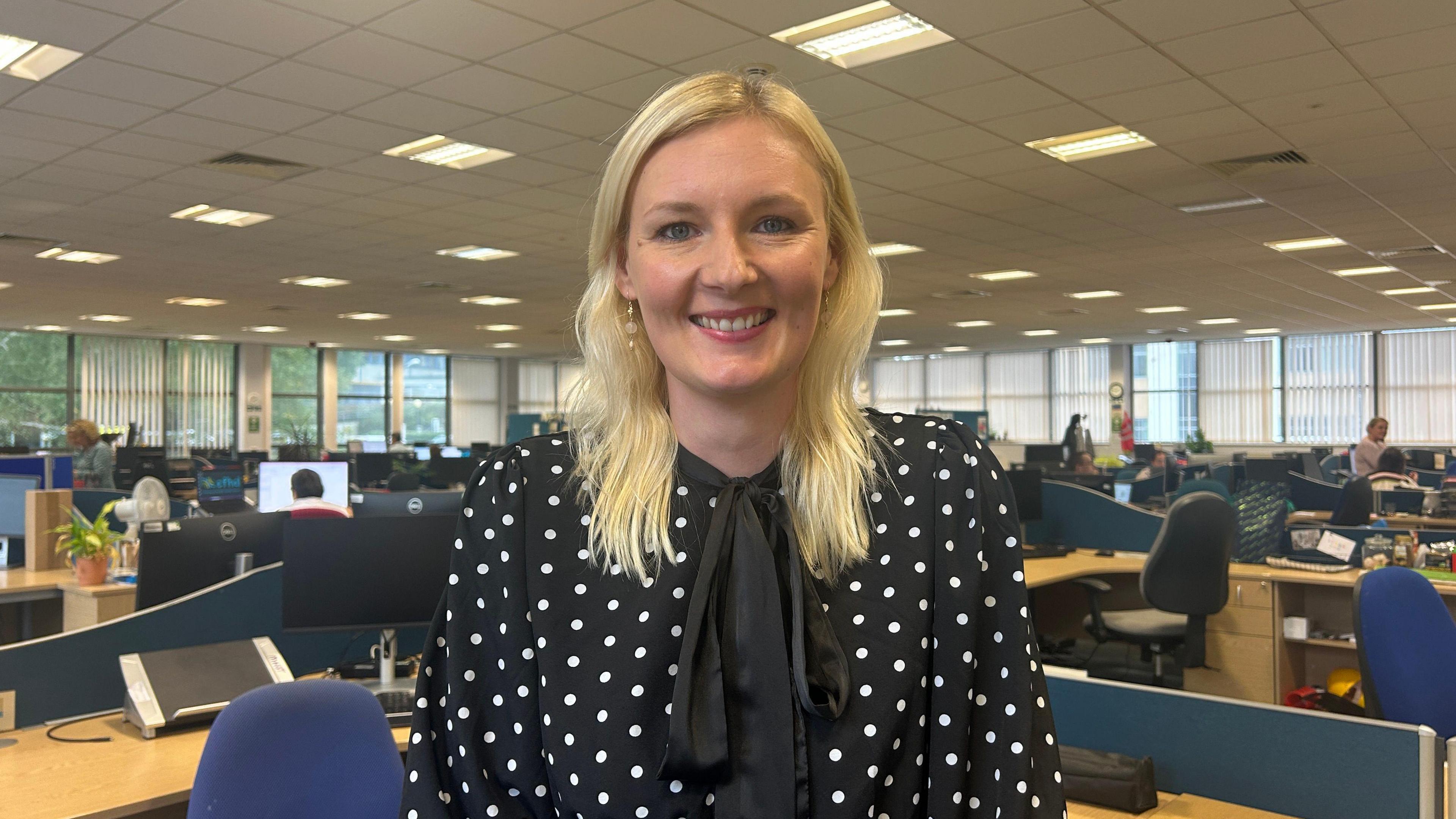 A woman with long blond hair standing in an office wearing a black spotty top