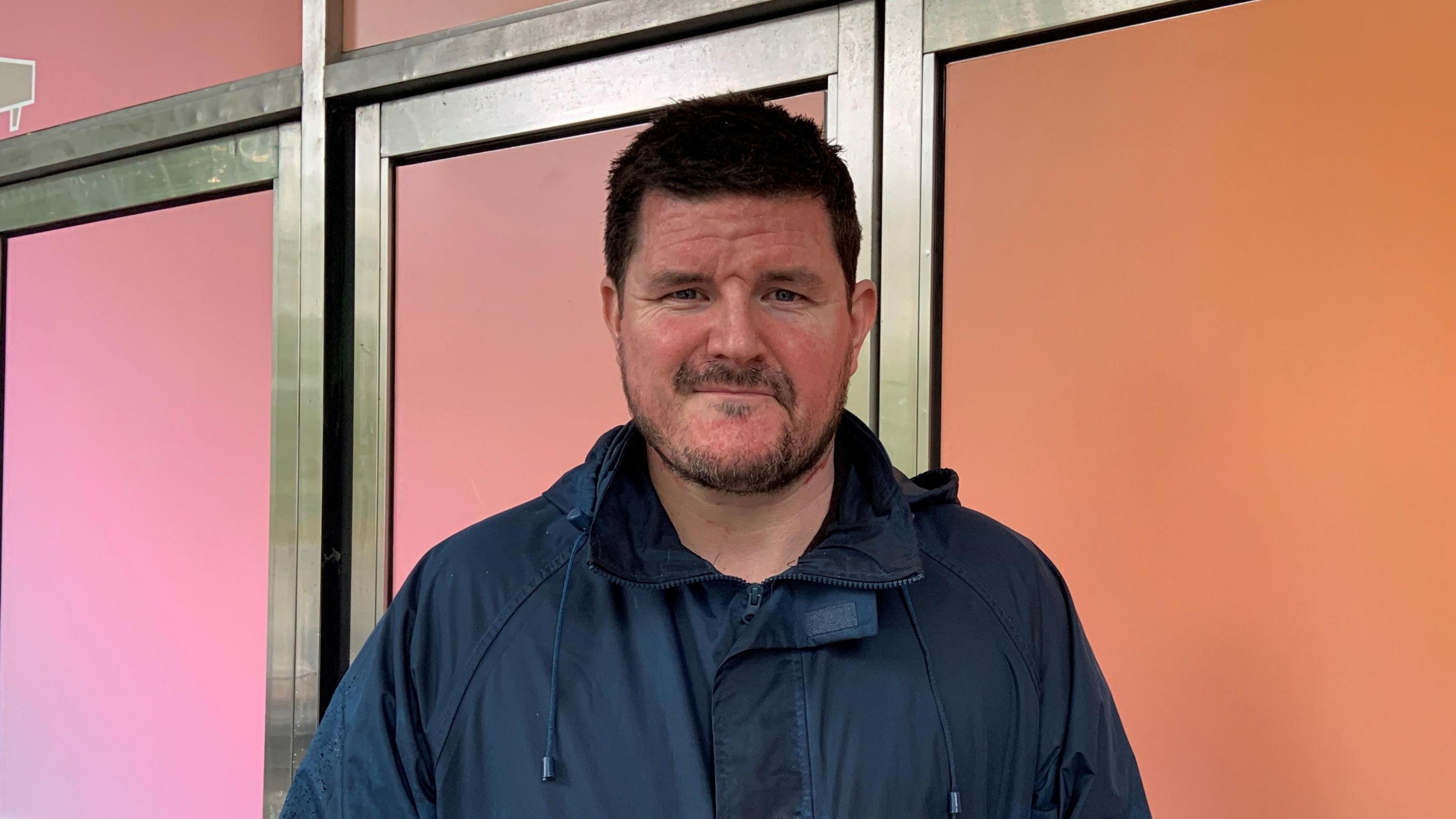 A brown haired male wearing a navy rain jack stares at the camera